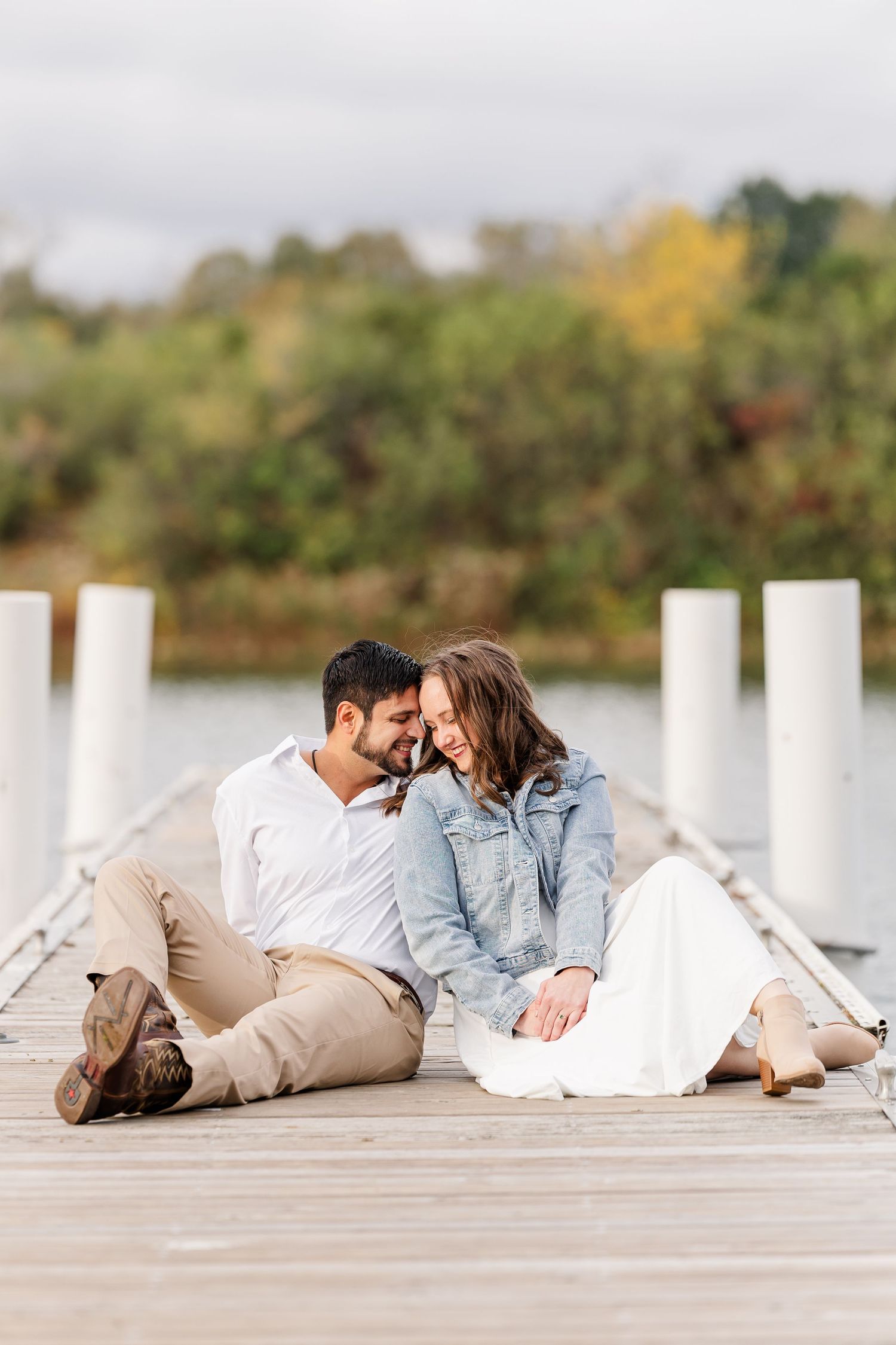 Smiling couple sitting close together on a wooden dock beside a calm lake with autumn trees during an outdoor engagement photo shoot.