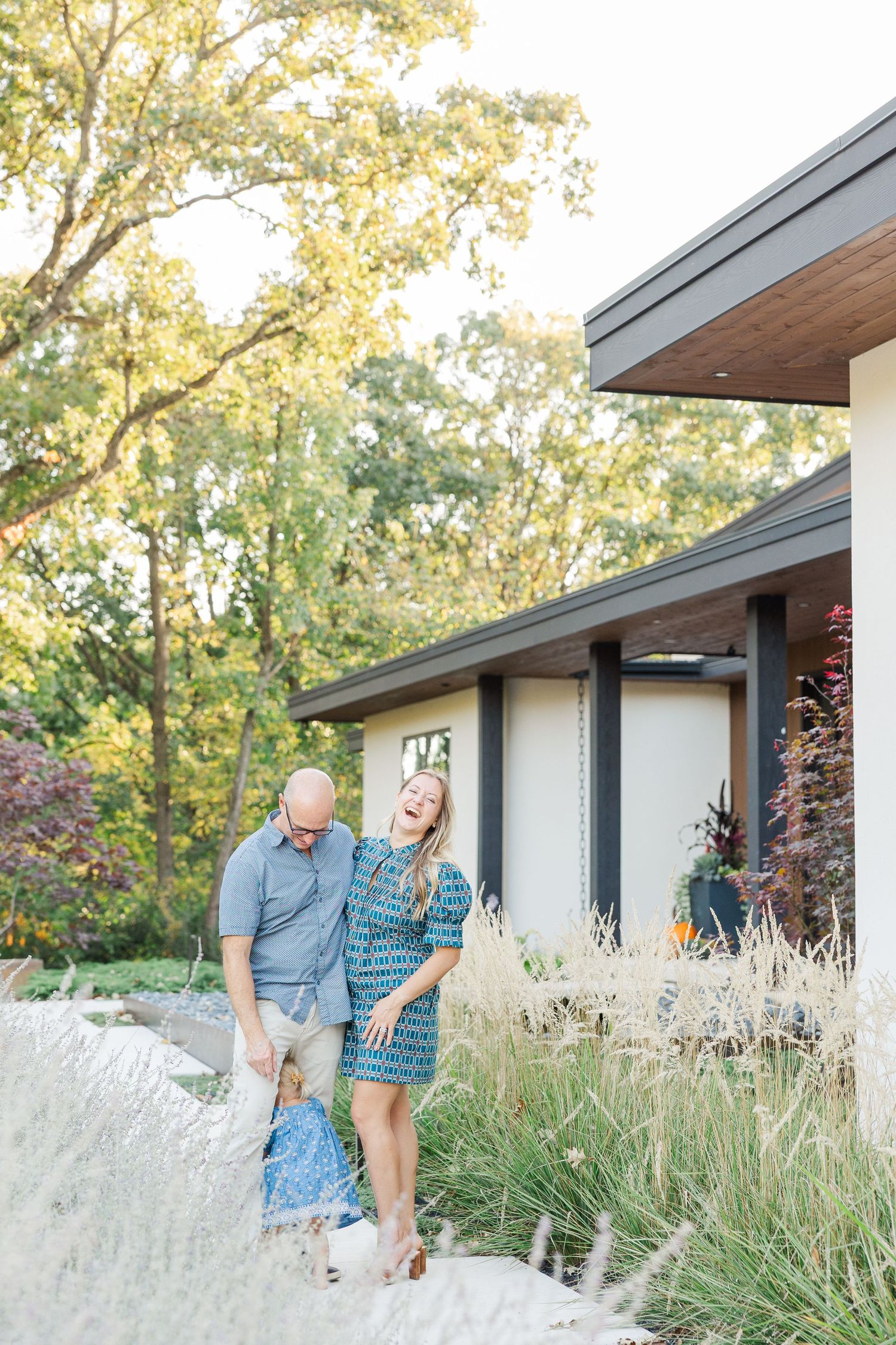 A couple shares a playful moment together in front of a modern house surrounded by ornamental grasses and trees.