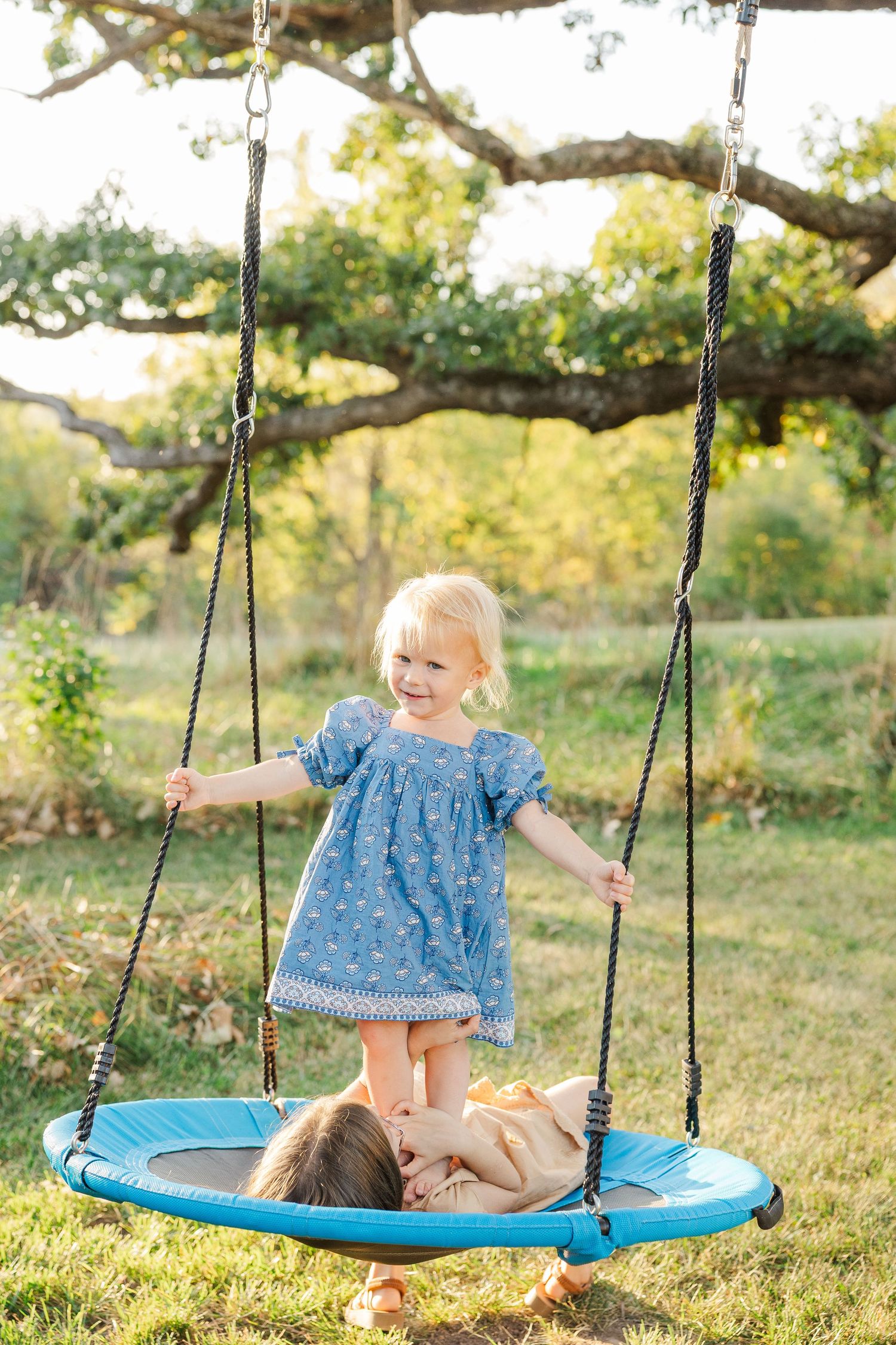 Children playing and laughing together on a blue disc tree swing in a sunny backyard setting at golden hour.