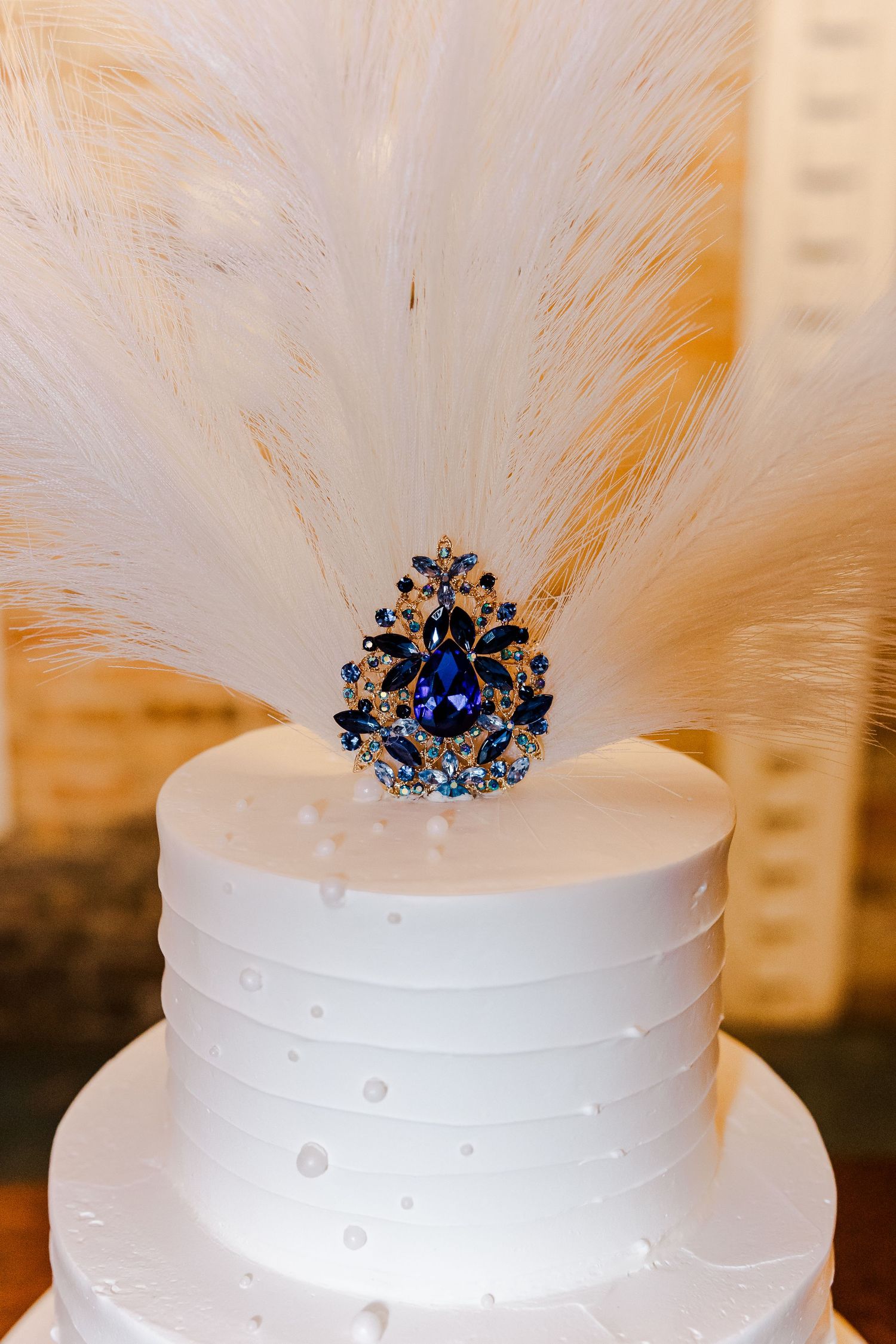 Elegant white wedding cake topped with blue jeweled brooch and white feather decoration.