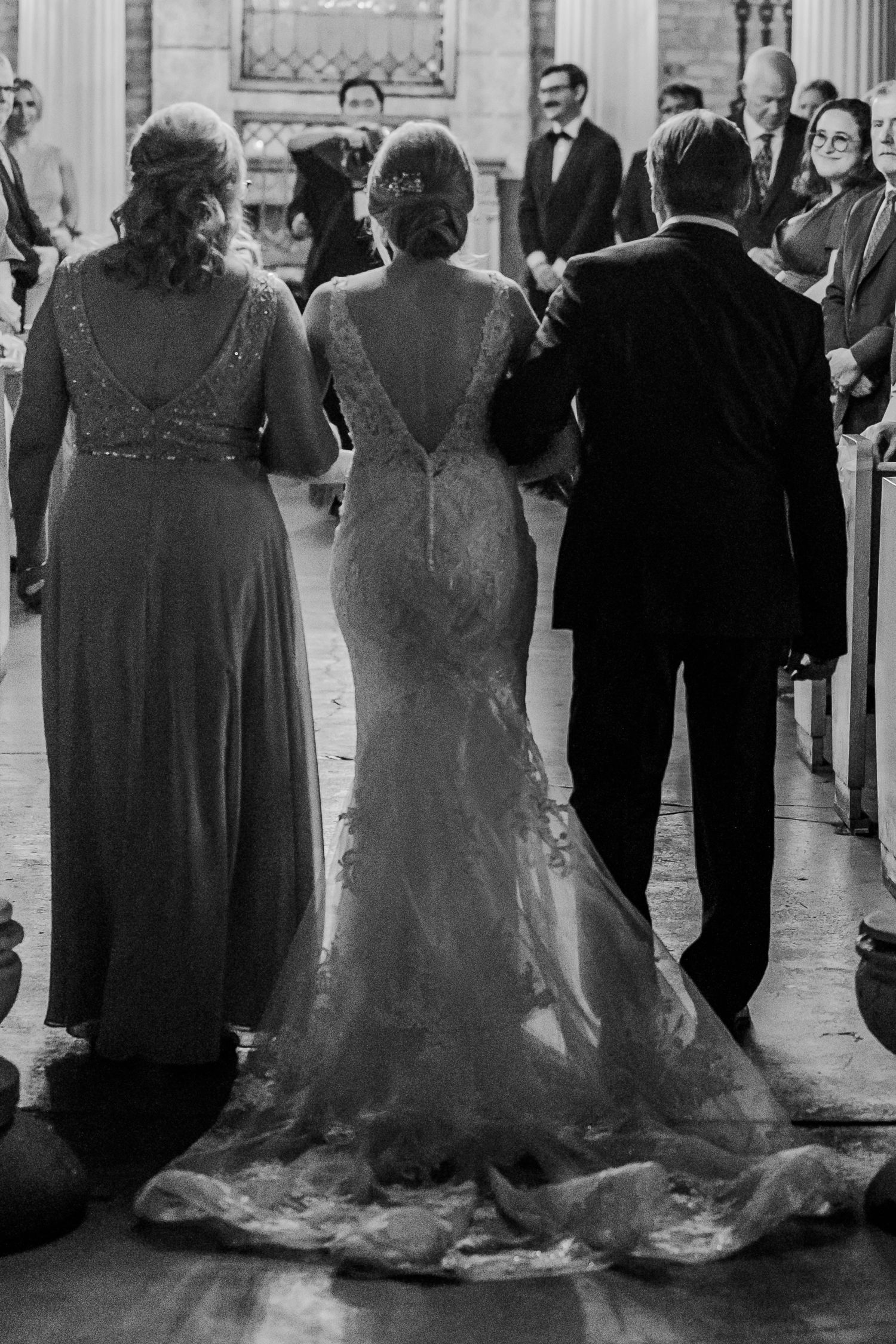 Back view of wedding party members in formal attire standing in elegant venue with flowing dress trains.
