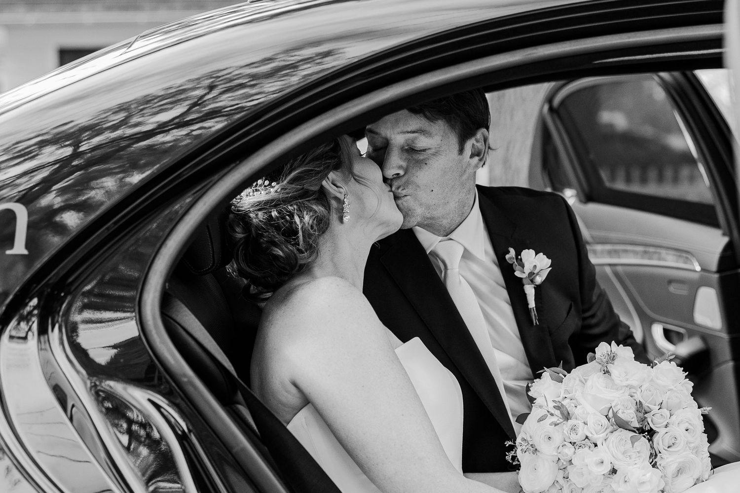 A romantic moment captured inside a luxury car on a wedding day with white floral bouquet visible.