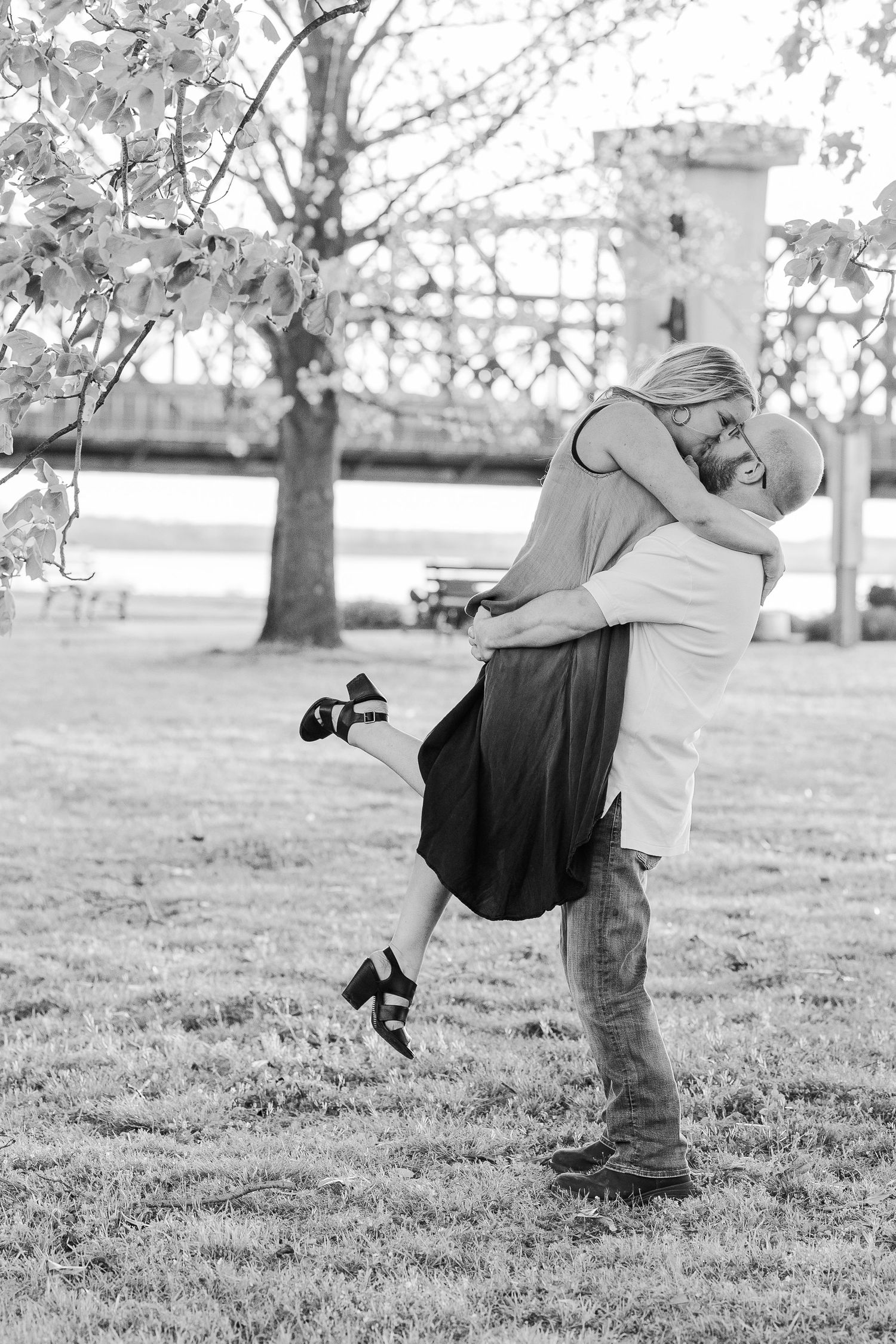 A black and white photo captures a romantic lift pose between a couple in a park setting.
