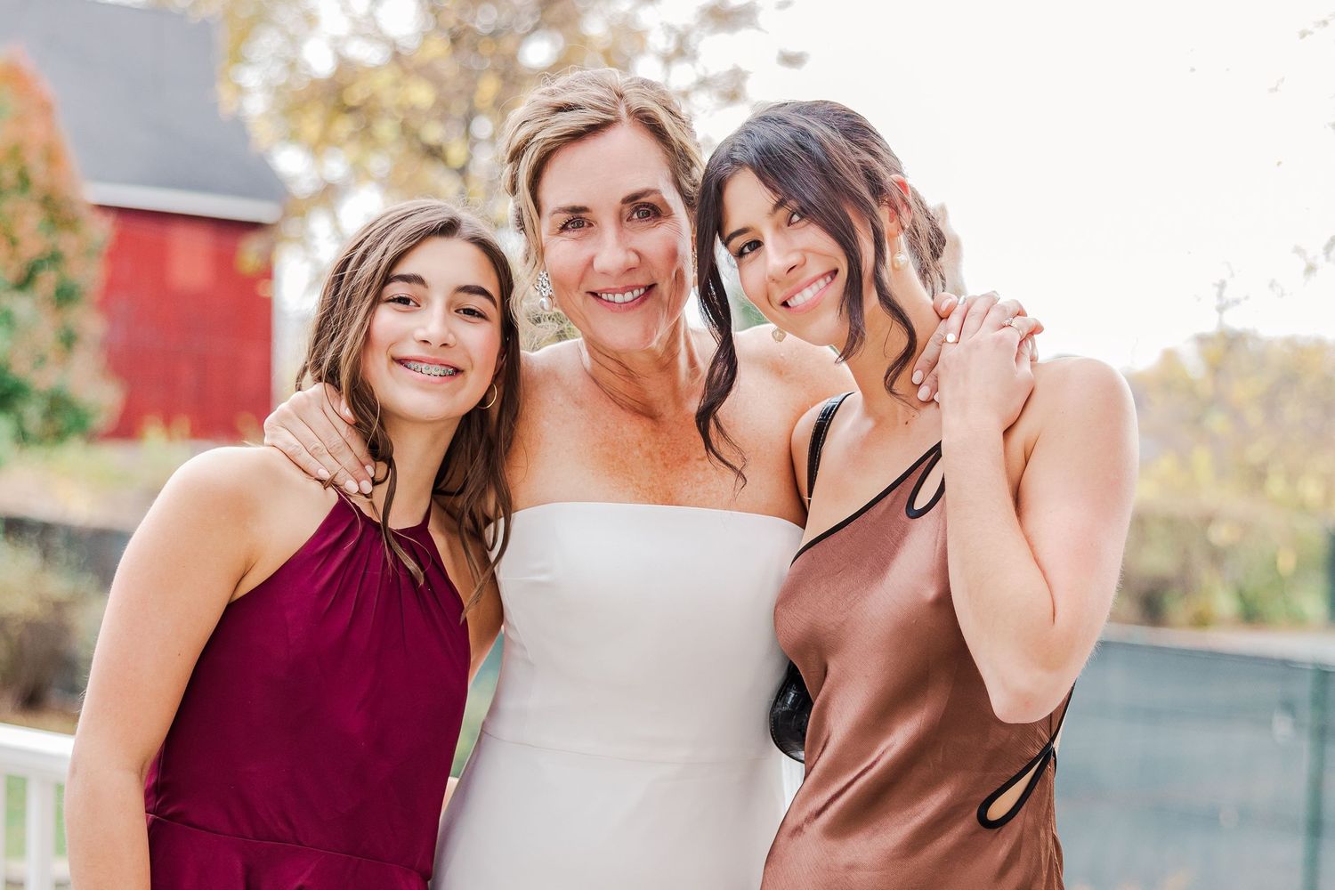 Three women in jewel-toned dresses pose together outdoors with a red barn in the background.