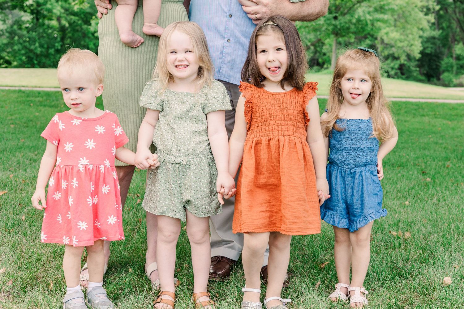 Four young children holding hands while wearing colorful summer dresses outdoors.