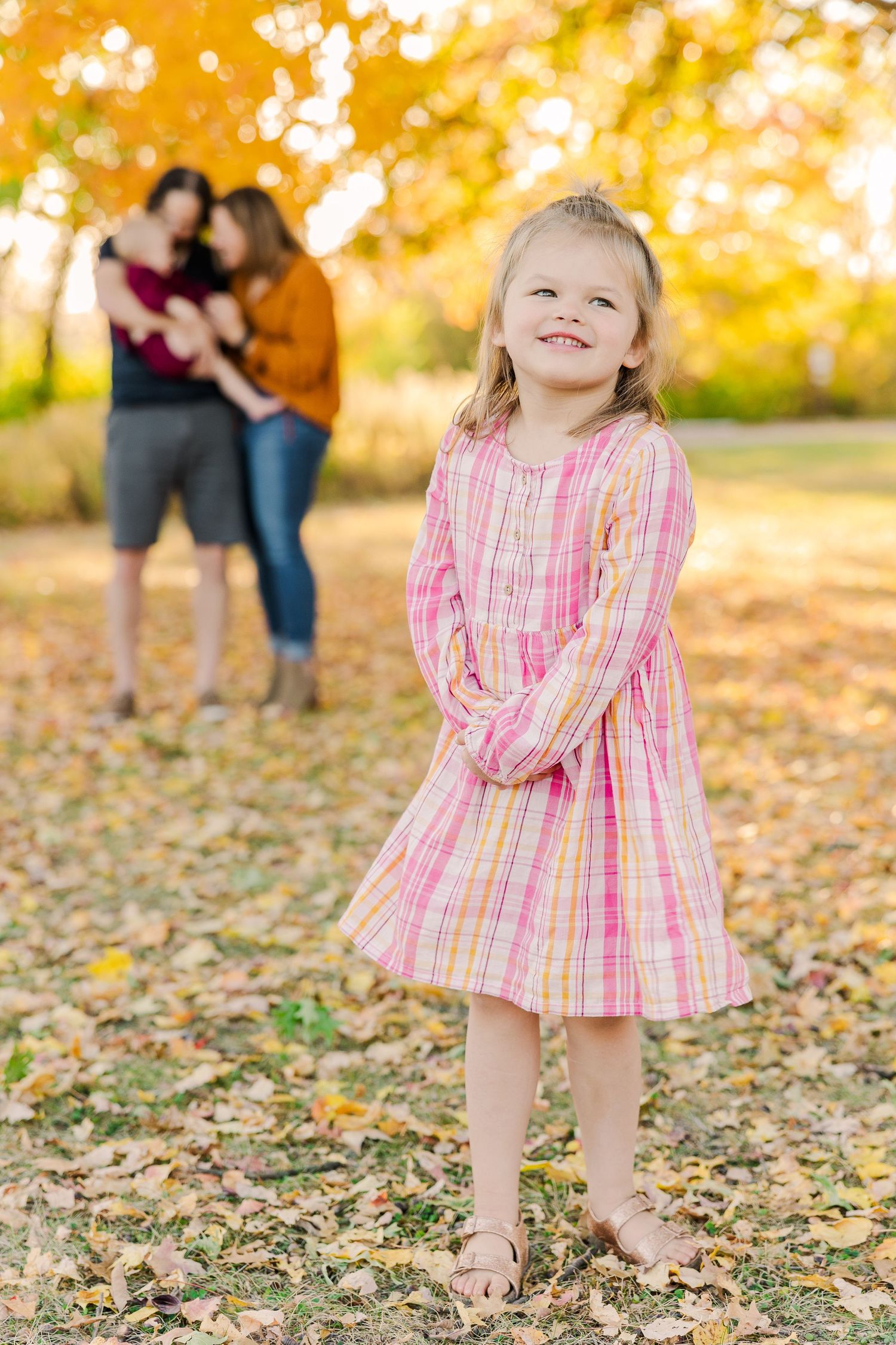 A child in a pink plaid dress smiles in autumn leaves with adults blurred in background.