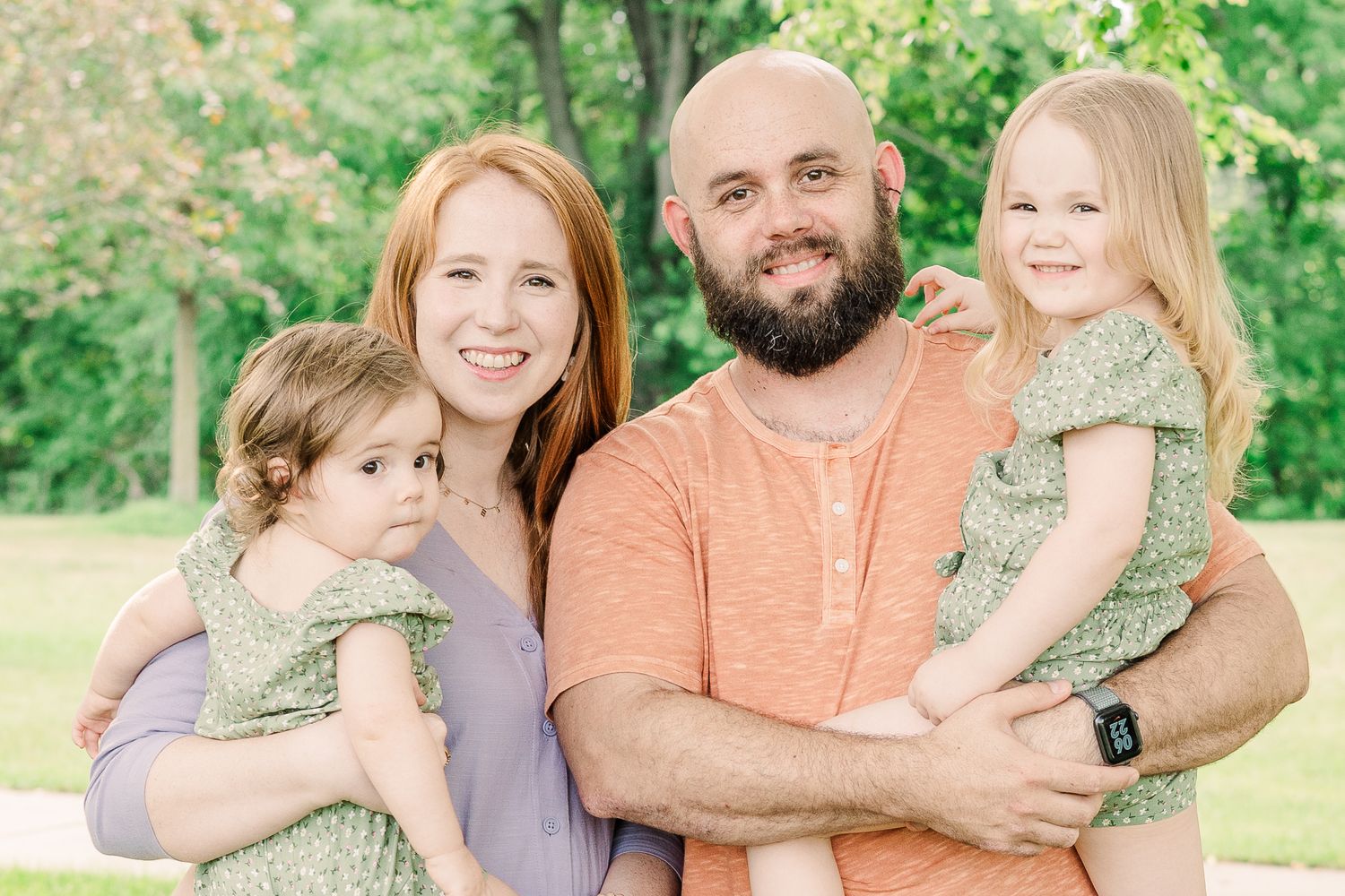 A loving family of four poses together outdoors against a green background on a sunny day.