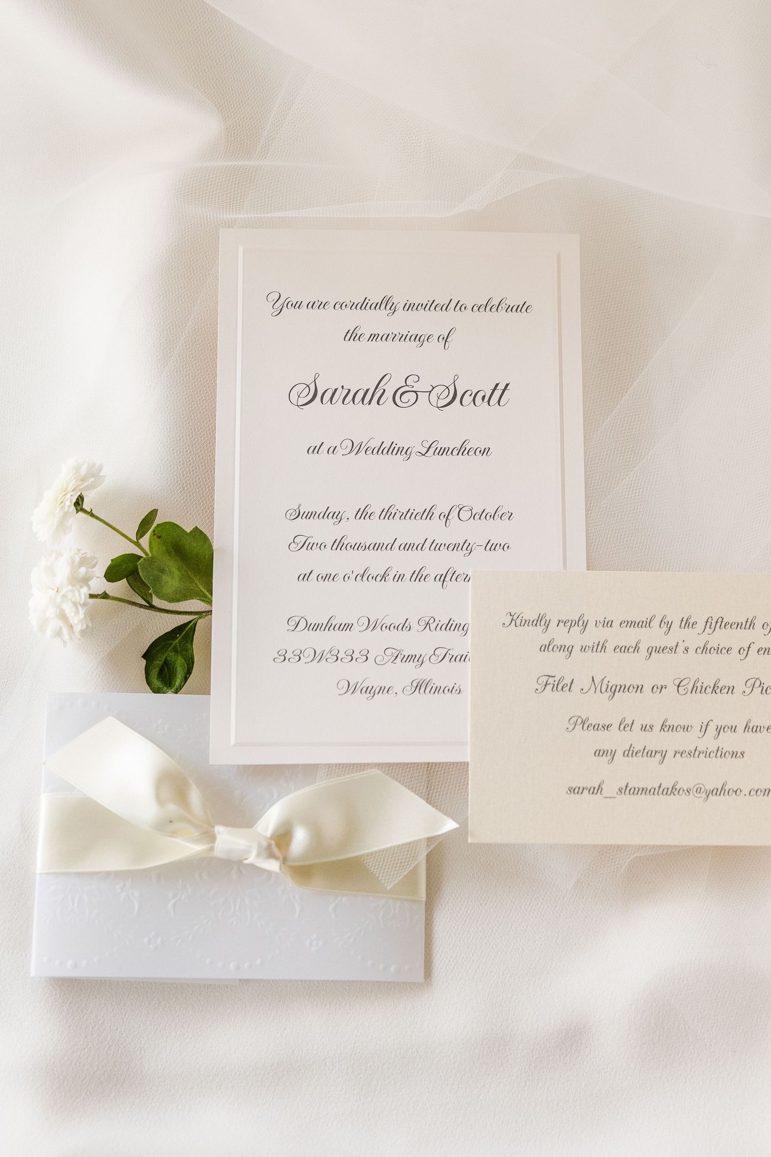 Elegant wedding invitation with white bow and floral accents on white fabric.