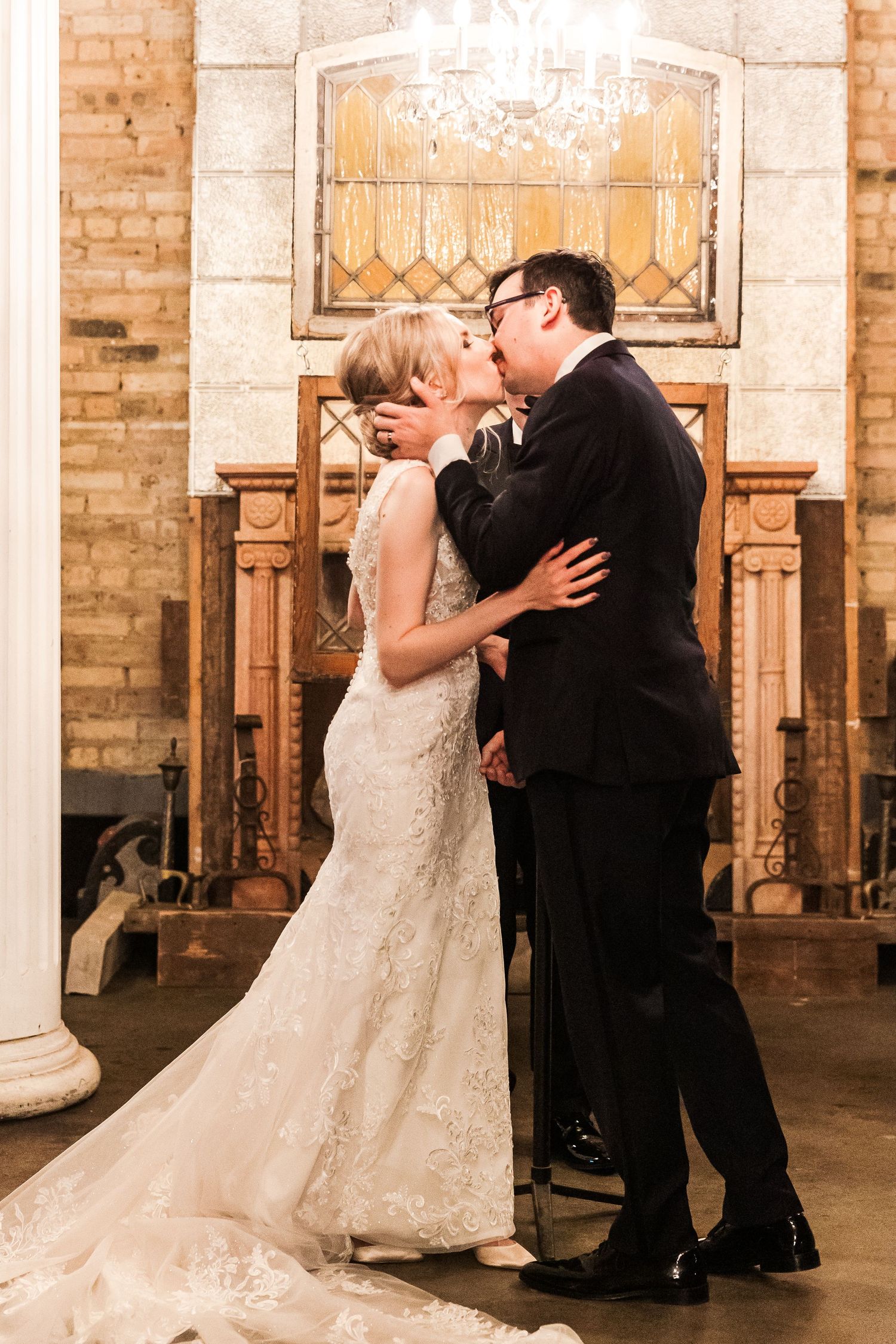 A wedding couple shares a romantic kiss under an ornate chandelier in front of decorative woodwork.