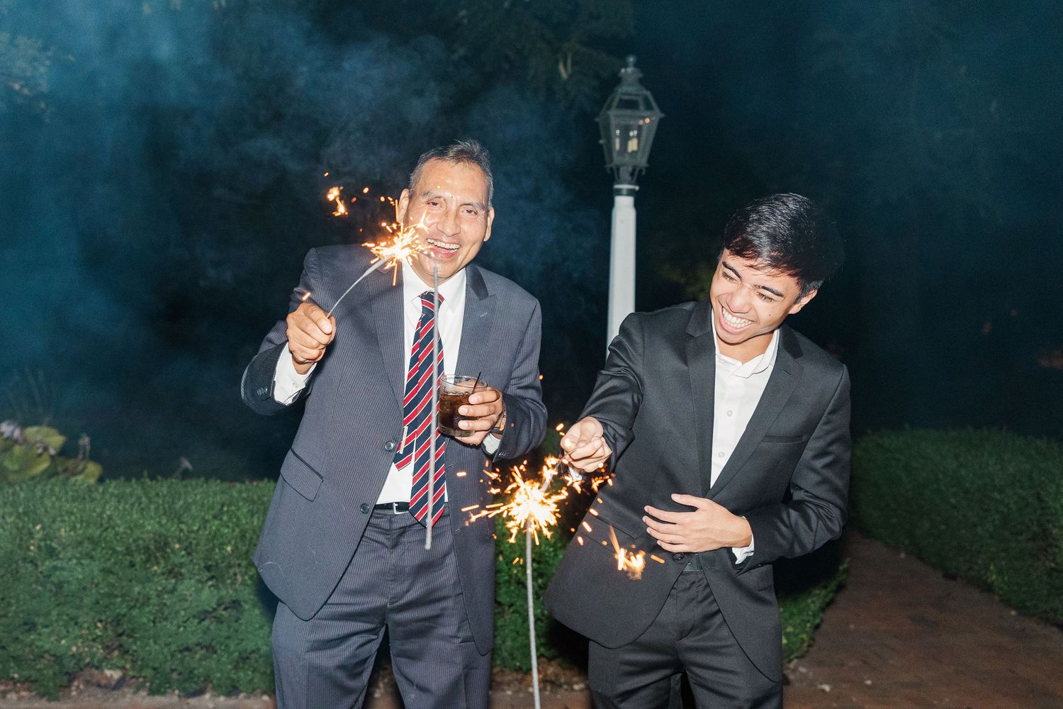 Two guests hold sparklers while celebrating outdoors at night near a decorative lamp post.