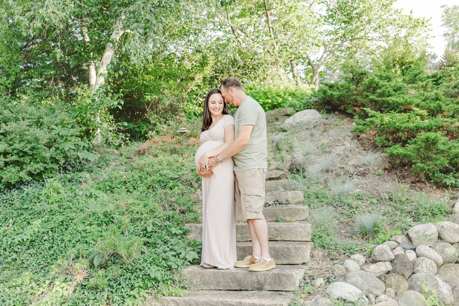 A couple stands together embracing on rustic stone steps surrounded by lush greenery on a sunny day.