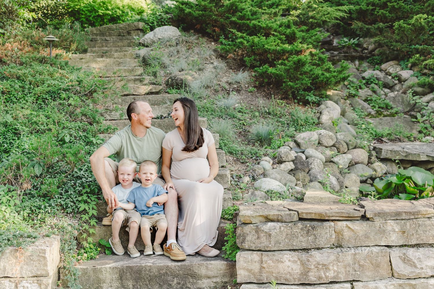 Family sits together on stone steps surrounded by lush green foliage during an outdoor photo session.