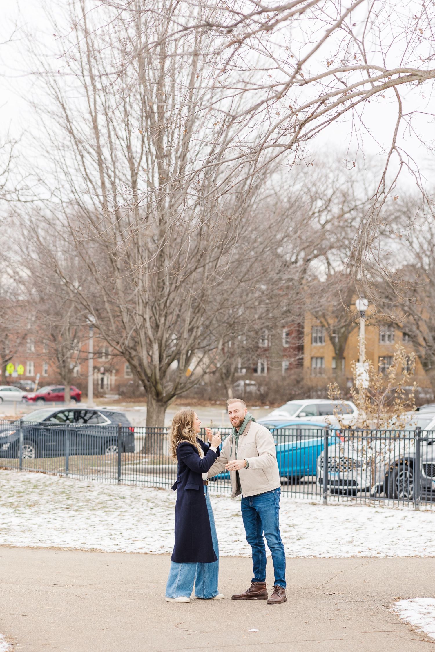 Man proposes to woman in snow-dusted park with bare trees and parked cars visible behind an iron fence in winter. Winnemac Park, Chicago