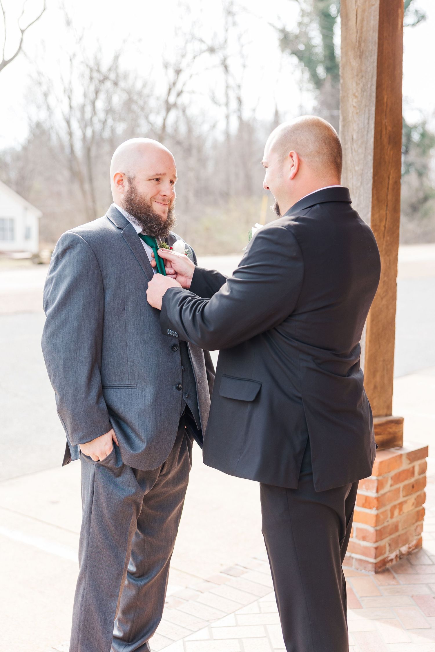 Two people in dark suits preparing formal attire on brick porch.