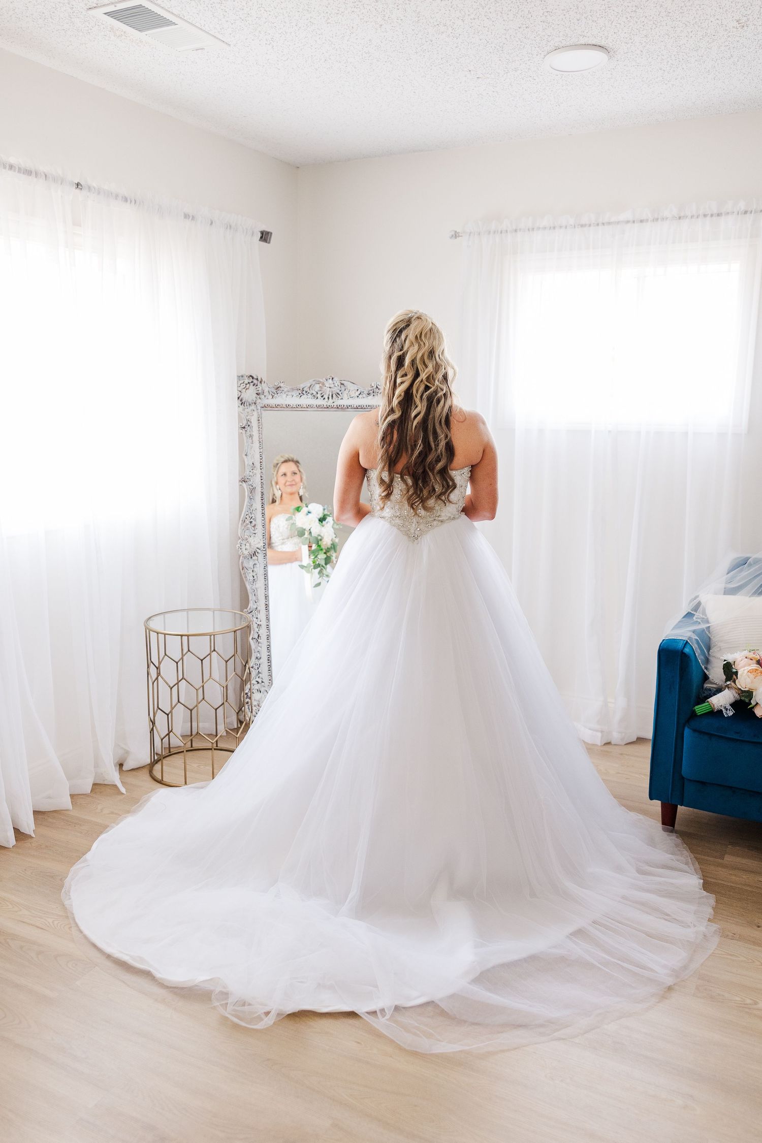 Back view of a princess ballgown wedding dress with flowing train in front of white curtains.