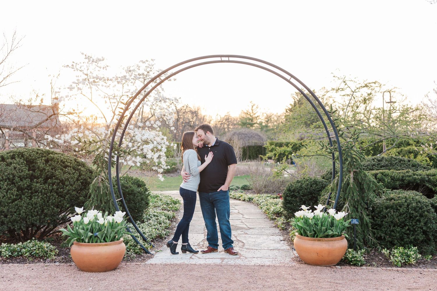 Happy couple posing beneath metal garden arch surrounded by manicured bushes and white tulips.