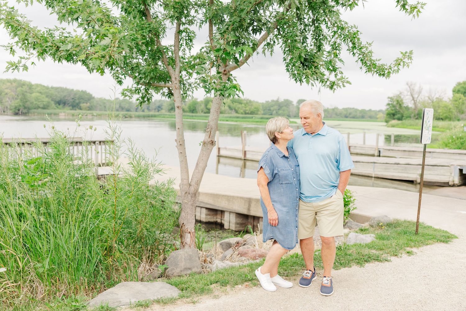 Two people stroll along a lakeside path near a wooden dock on a bright summer day.