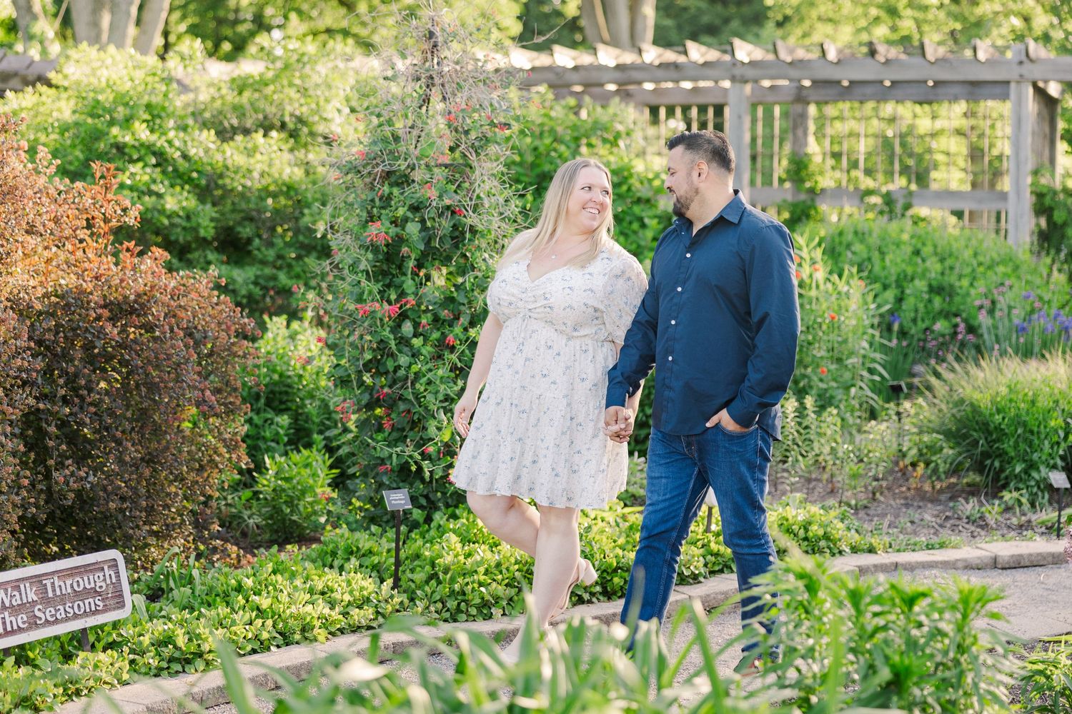 A romantic couple walks through a garden setting with blooming flowers and greenery.