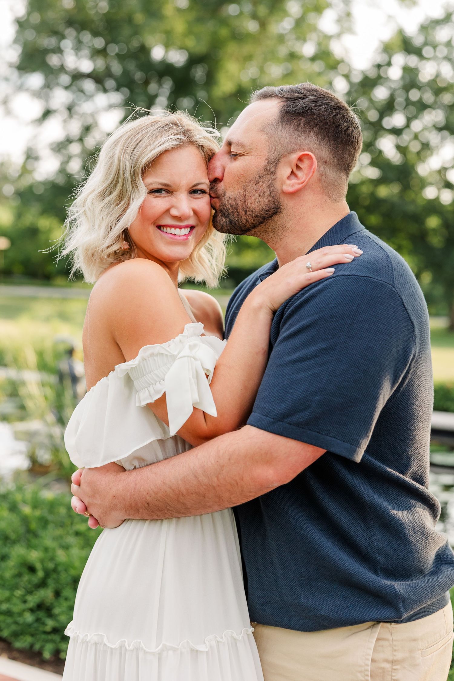 A man in a navy shirt kisses a smiling blonde woman in a white dress in a lush garden setting.