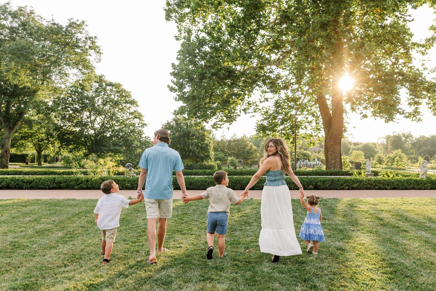Family of five walking hand-in-hand through a lush green park at golden hour, with sunlight filtering through tall trees.