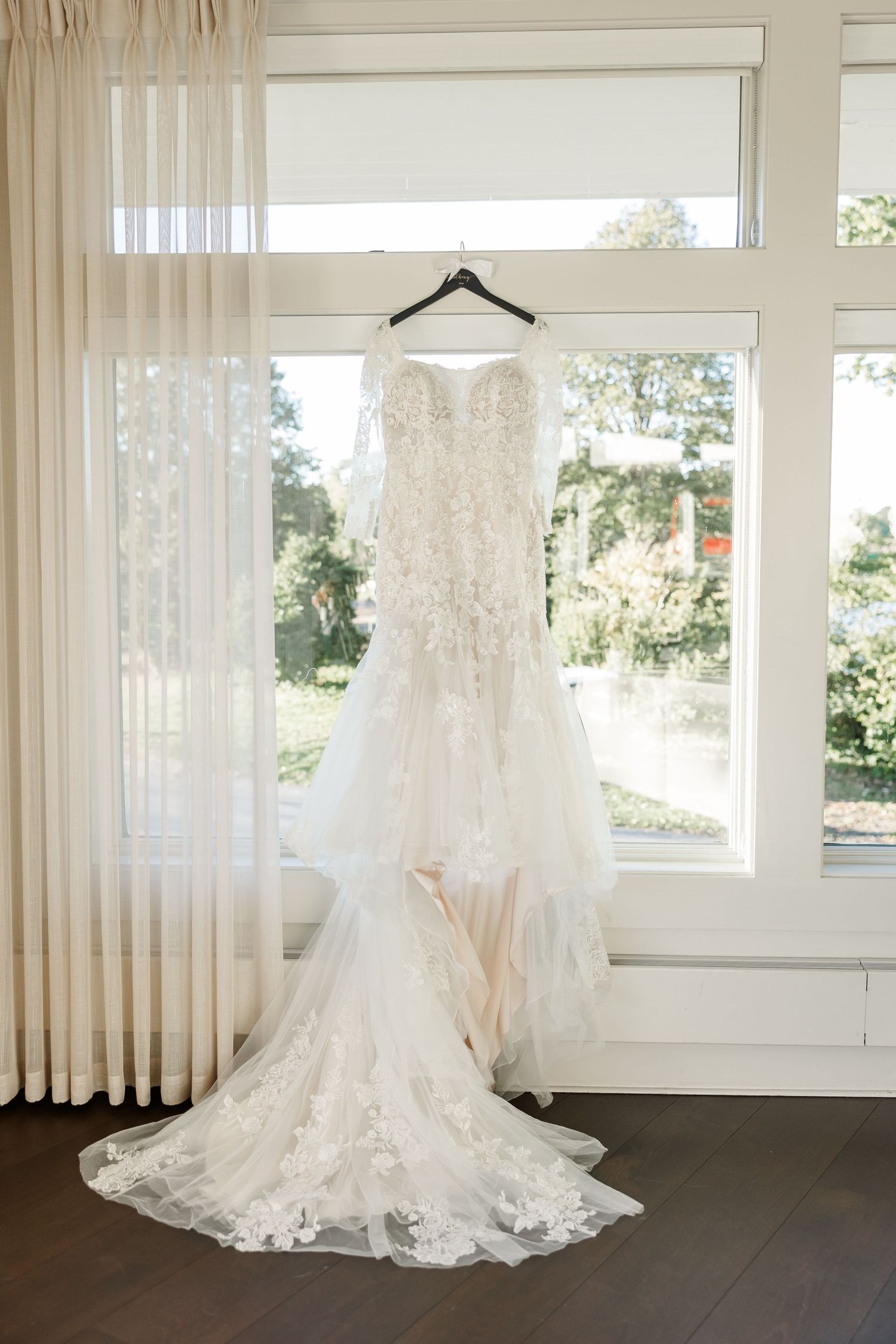Ivory lace wedding gown with floral appliqués and a long tulle train hanging on a hanger in front of tall bright windows.