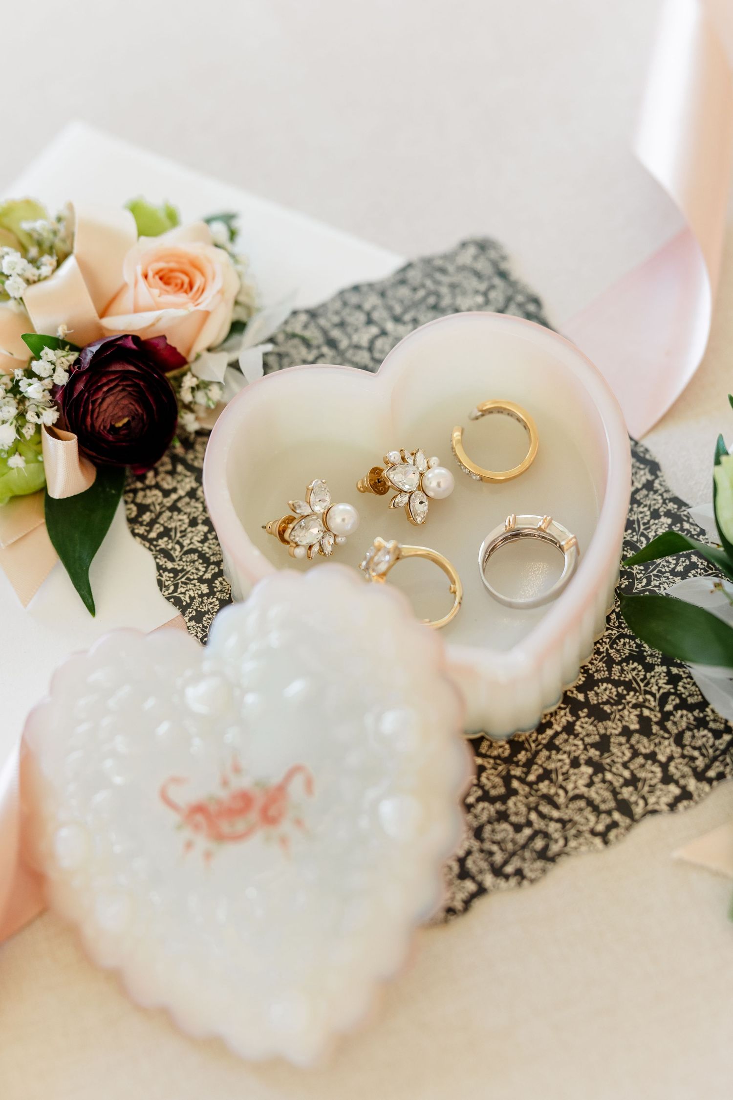 Heart-shaped milk glass ring dish holding gold wedding bands and pearl earrings, beside a lid and a peach rose boutonniere.