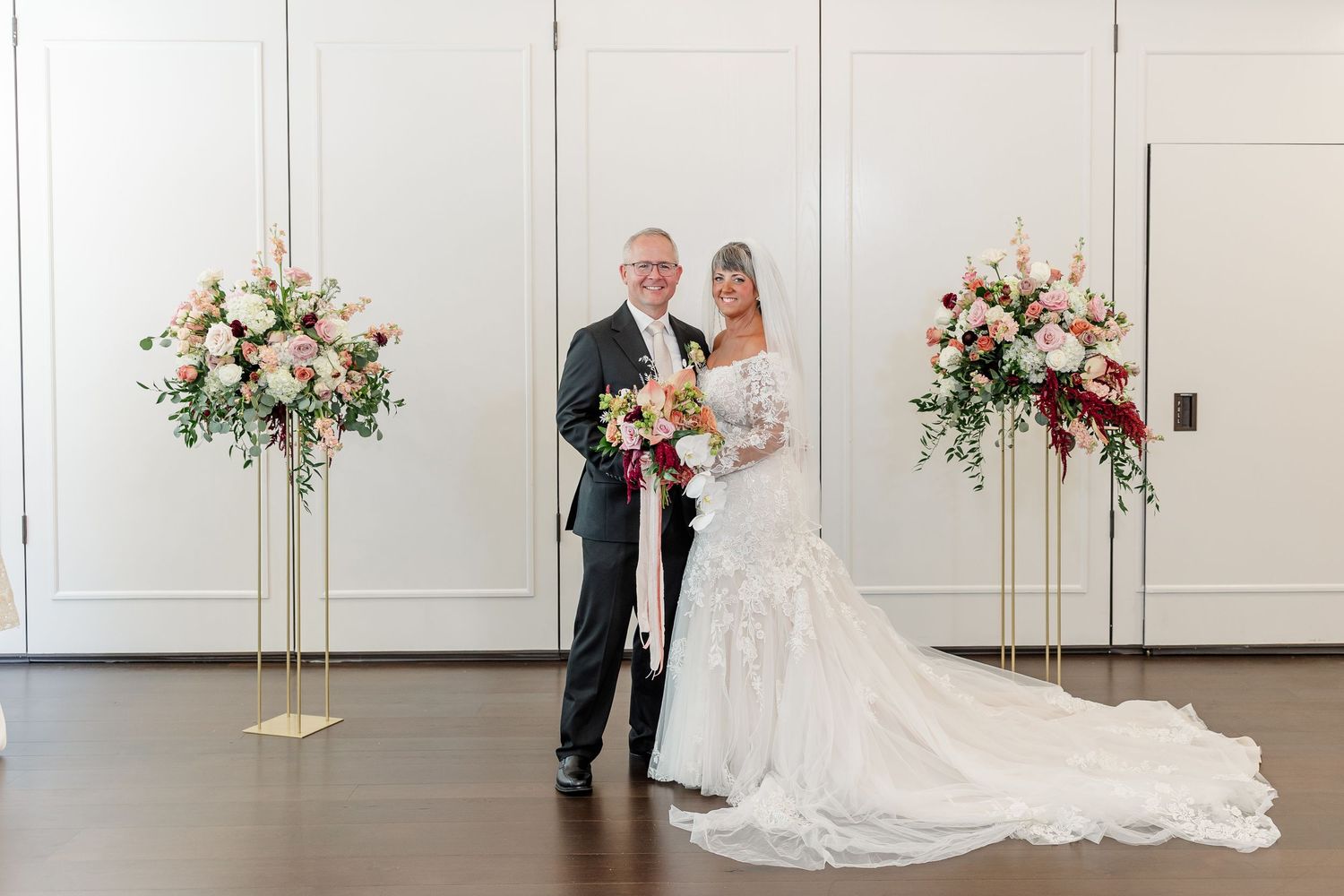 Bride in a lace wedding gown with a long train holds a bouquet beside her groom in a dark suit, flanked by tall pink floral arrangements.