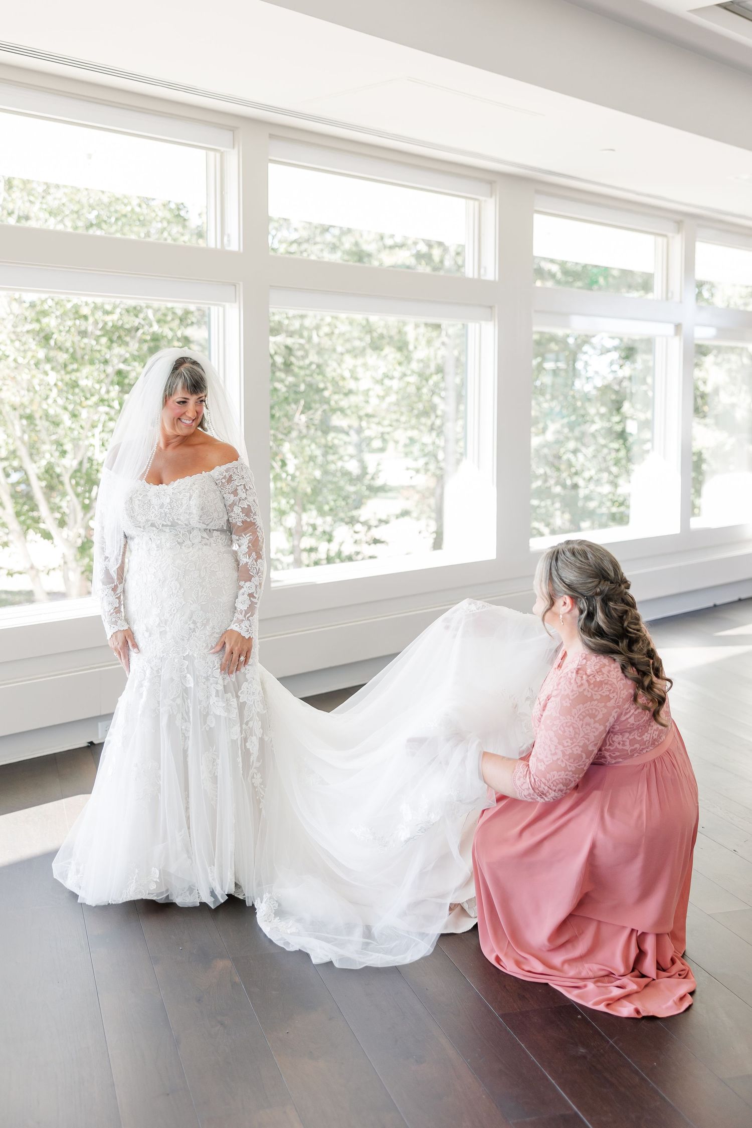 Bride in a lace off-the-shoulder wedding gown and veil stands in a bright room as a woman in a pink dress arranges her long train.