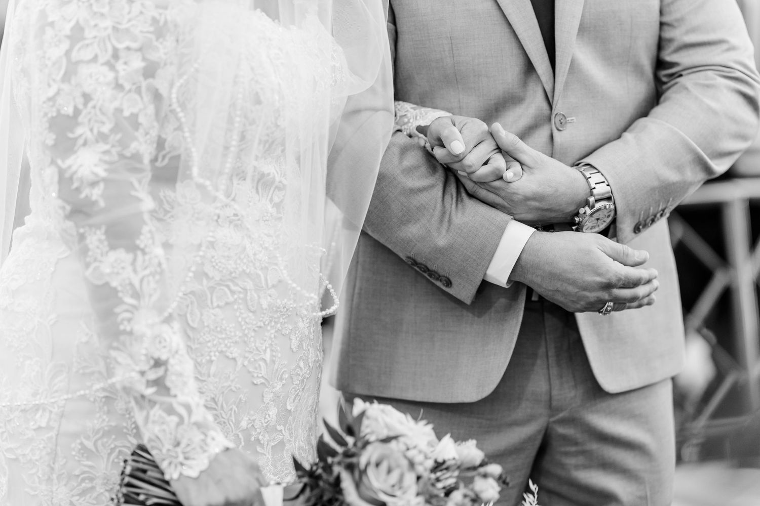 Black and white photo of a bride in a lace veil linking arms with a groom in a suit, holding a bouquet of roses at a wedding ceremony.