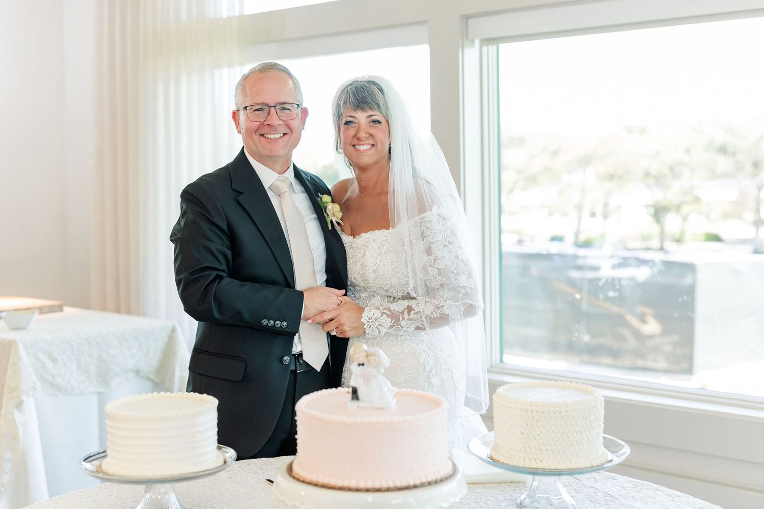 A smiling bride and groom pose together holding hands beside a pink wedding cake and two white tiered cakes by a bright window.