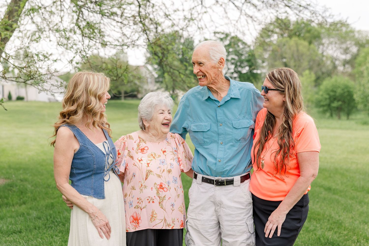 Two adult daughters laughing joyfully with their elderly parents standing together in a lush green outdoor setting.