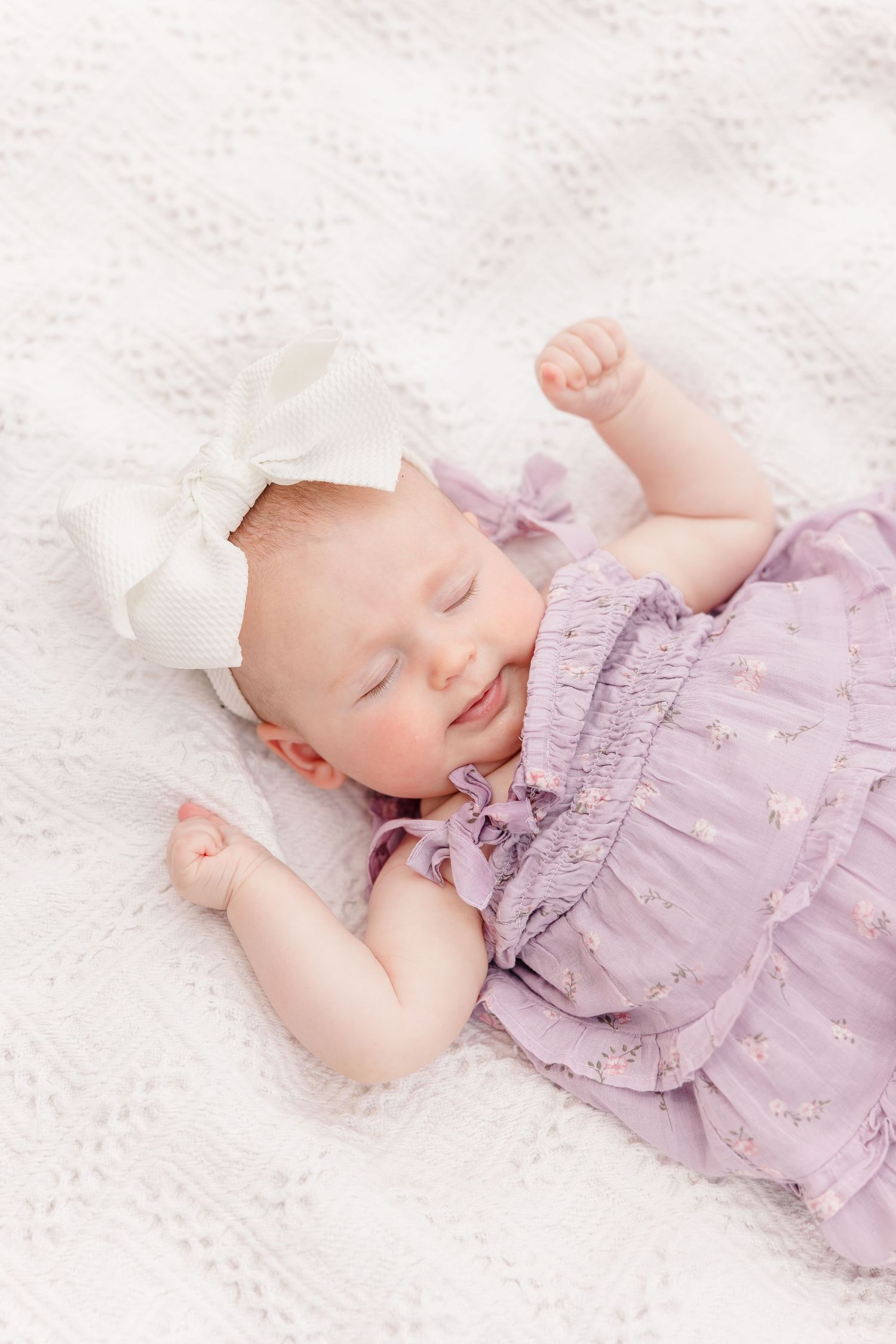 A peaceful newborn baby girl in a lavender ruffle dress and white bow sleeps on a white blanket.