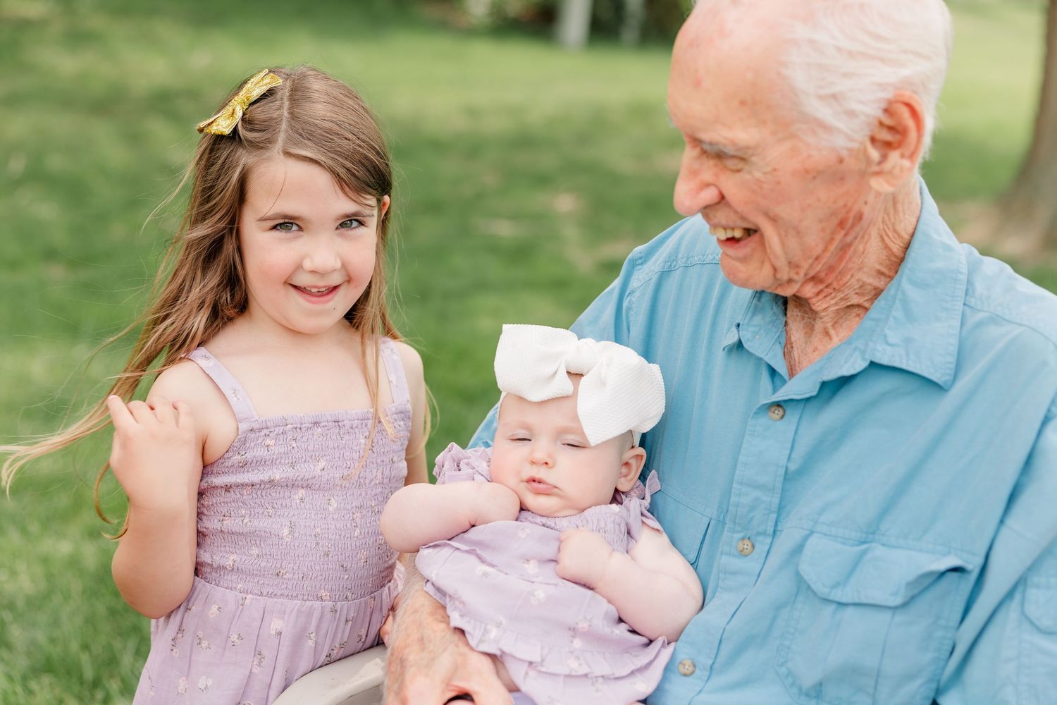 Elderly grandfather smiling warmly while holding a newborn baby alongside a young granddaughter in a purple dress.