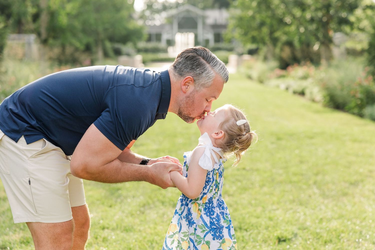 A grandfather lovingly kisses a toddler girl in a floral dress on a lush green lawn.
