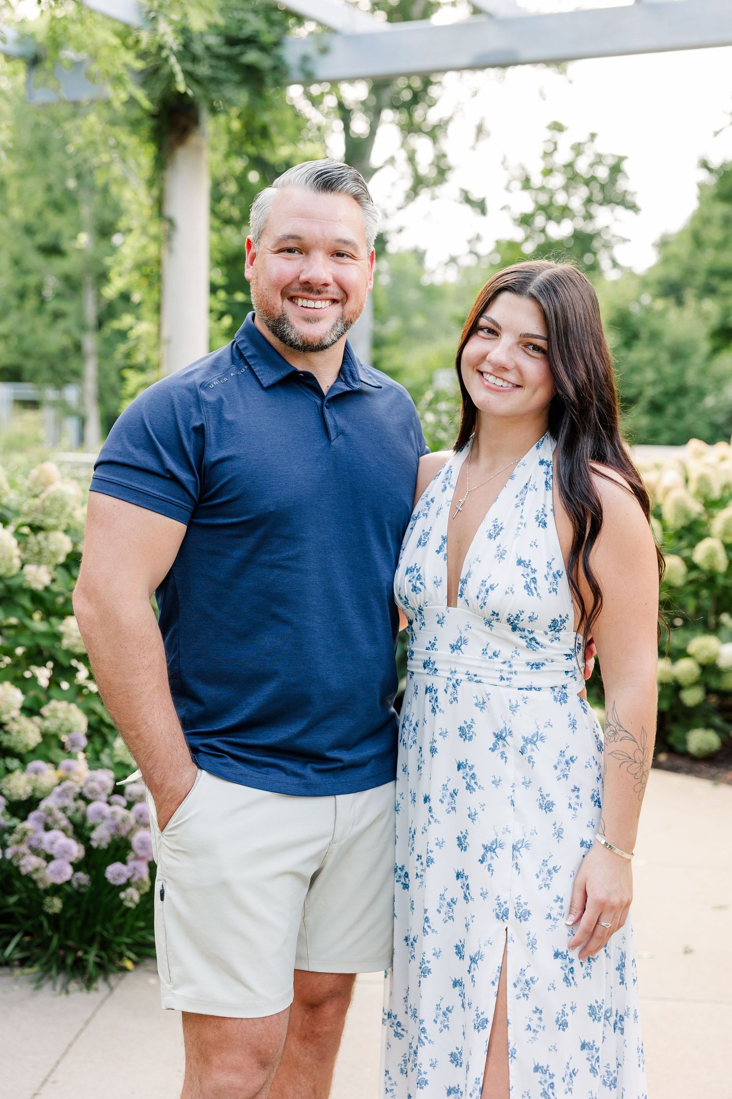 Smiling man in navy polo and young woman in blue floral dress posing among hydrangeas.
