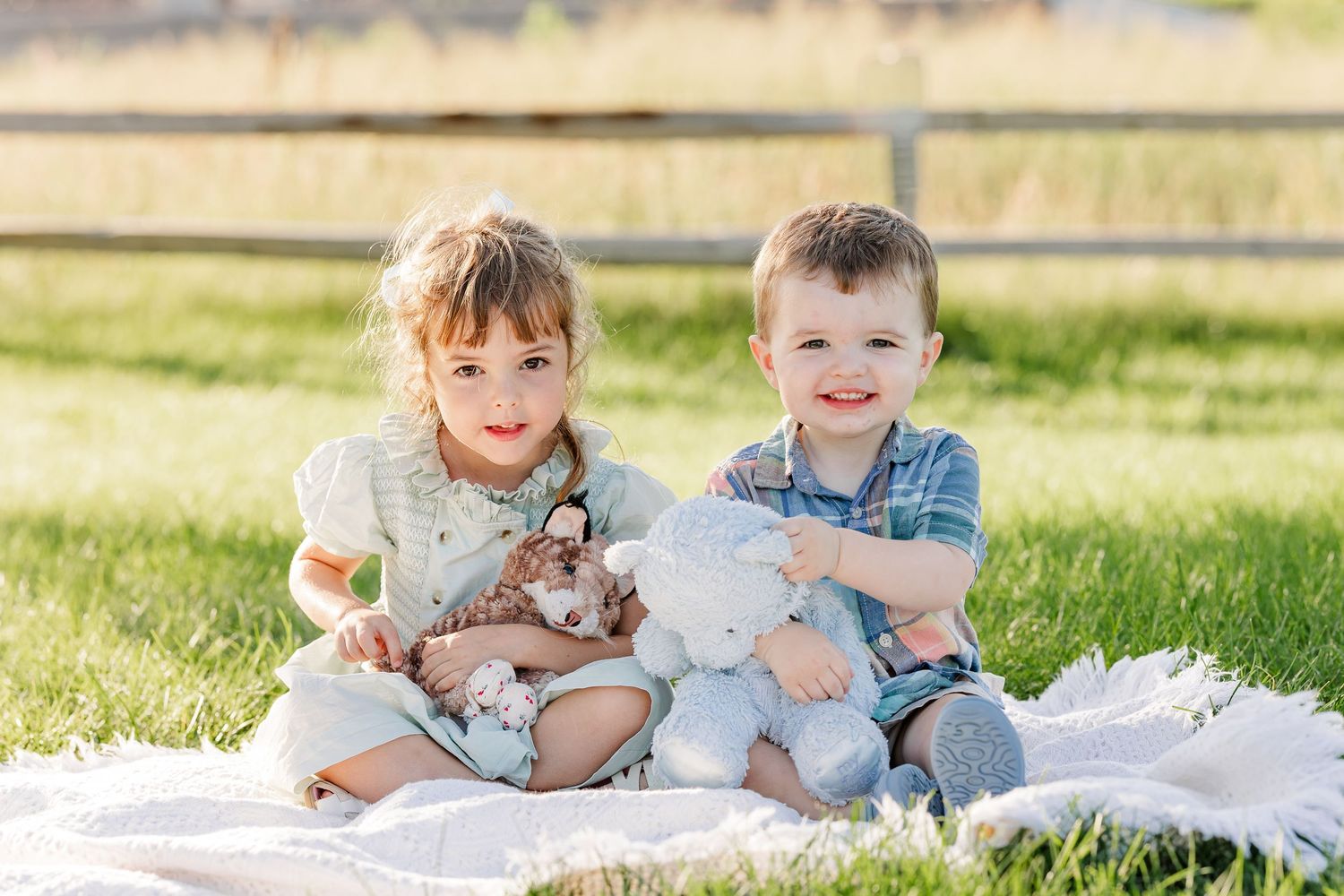 Two young children sitting on a white blanket in a sunny park, smiling and holding stuffed animal toys.