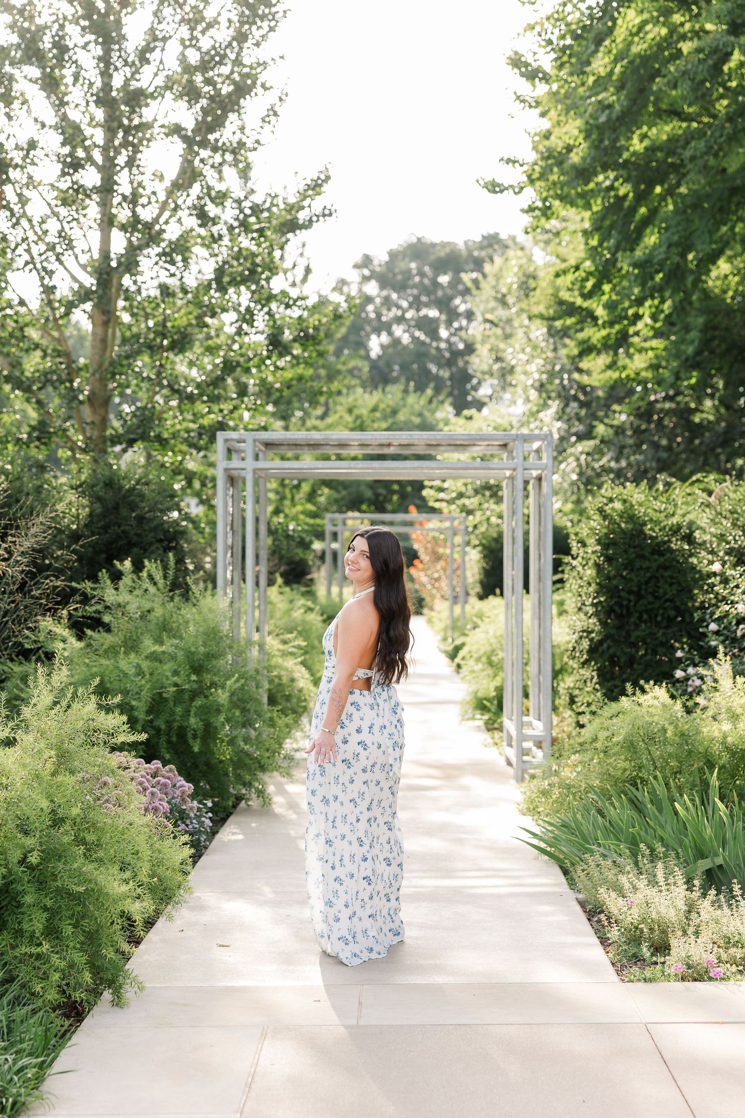 Young woman in white floral maxi dress walking along garden pathway under pergola surrounded by lush greenery.
