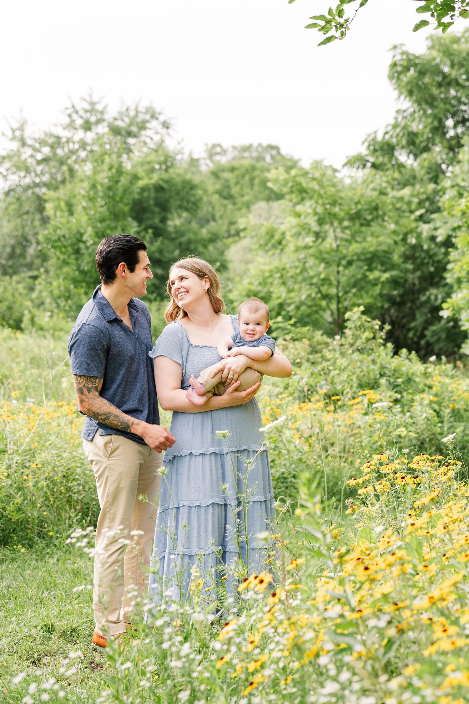 A happy young family stands together in a blooming wildflower meadow, mother holding their infant baby.