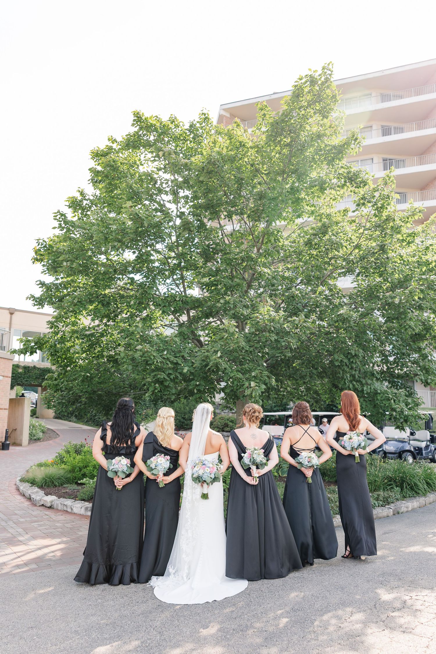 Bride in white gown with veil stands between bridesmaids in black dresses showing bouquets from behind.
