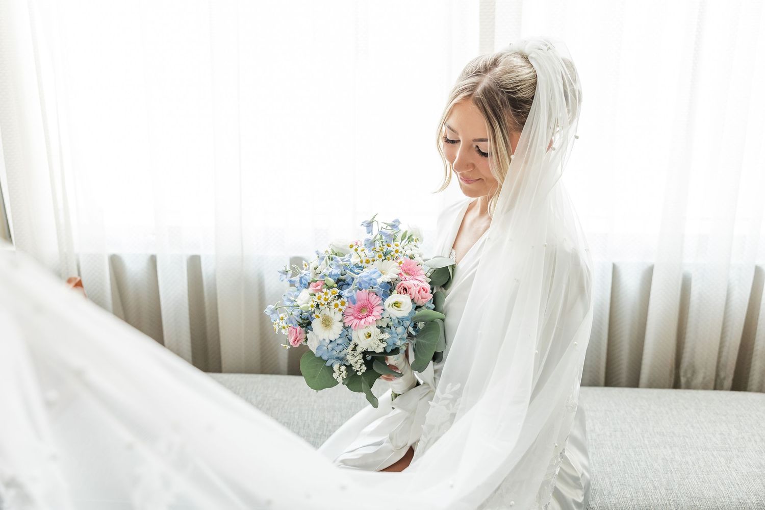 Bride in white wedding dress and veil holds pastel pink and blue floral bouquet while seated near bright window.
