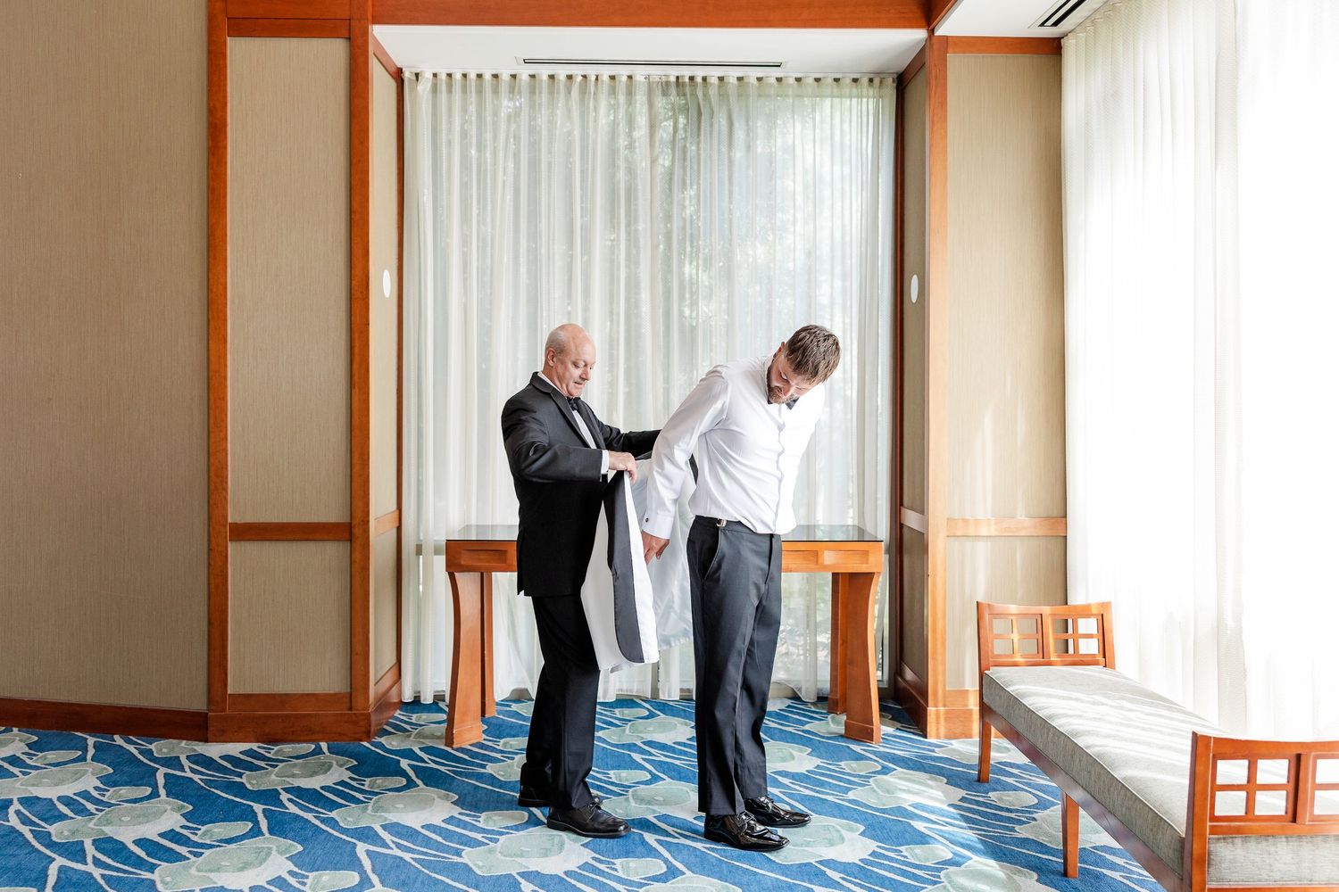 Father helping groom with jacket in hotel room with blue patterned carpet before wedding.