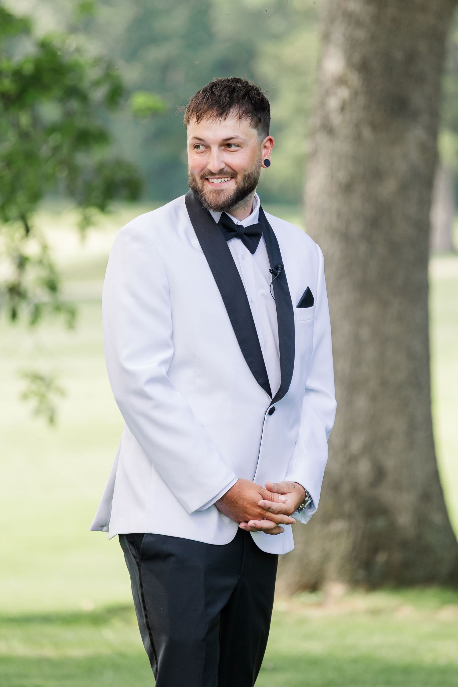 Smiling groom in white tuxedo jacket with black lapels standing outdoors near trees.