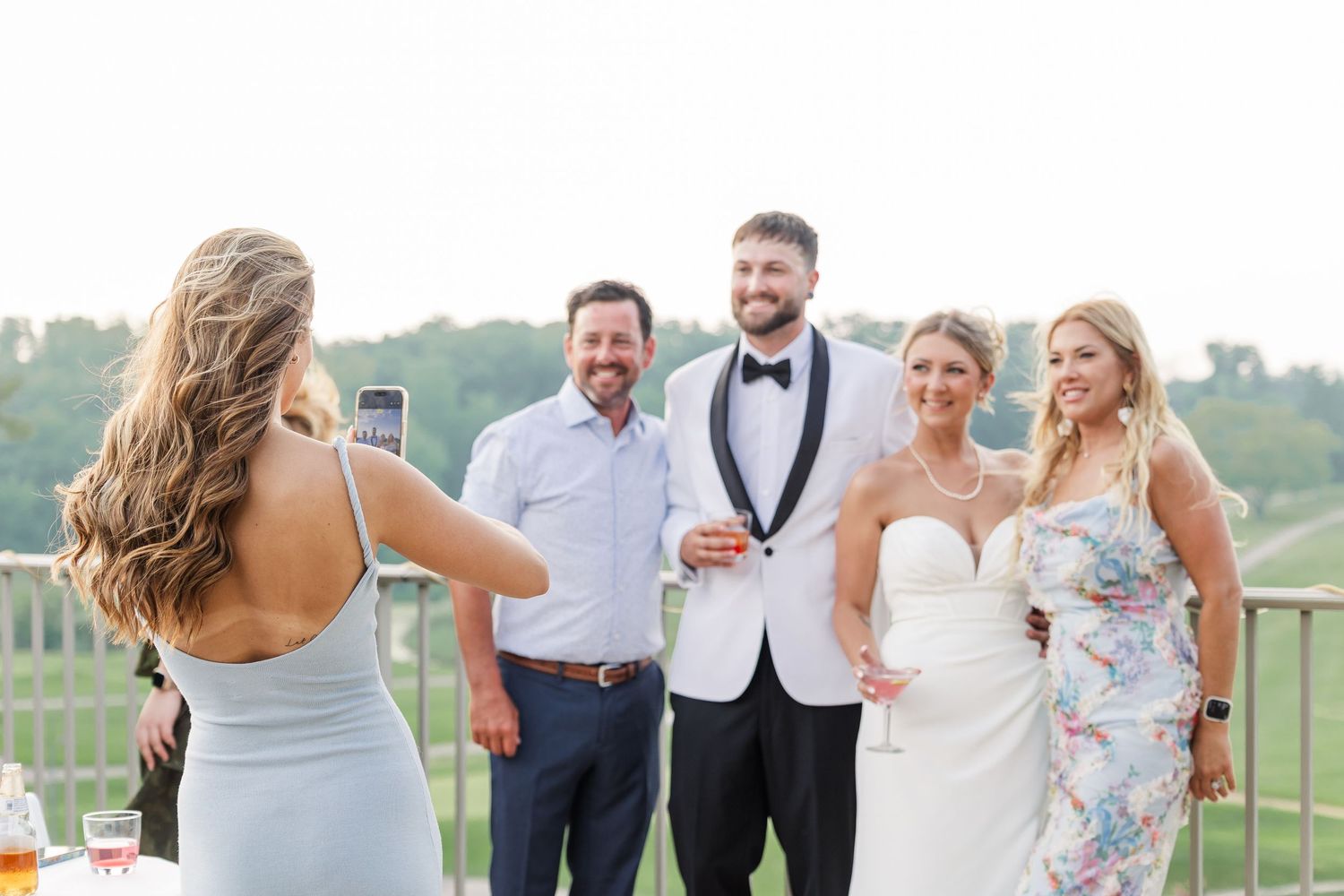 Wedding photographer capturing bride and groom posing with guests on an outdoor venue deck at sunset.