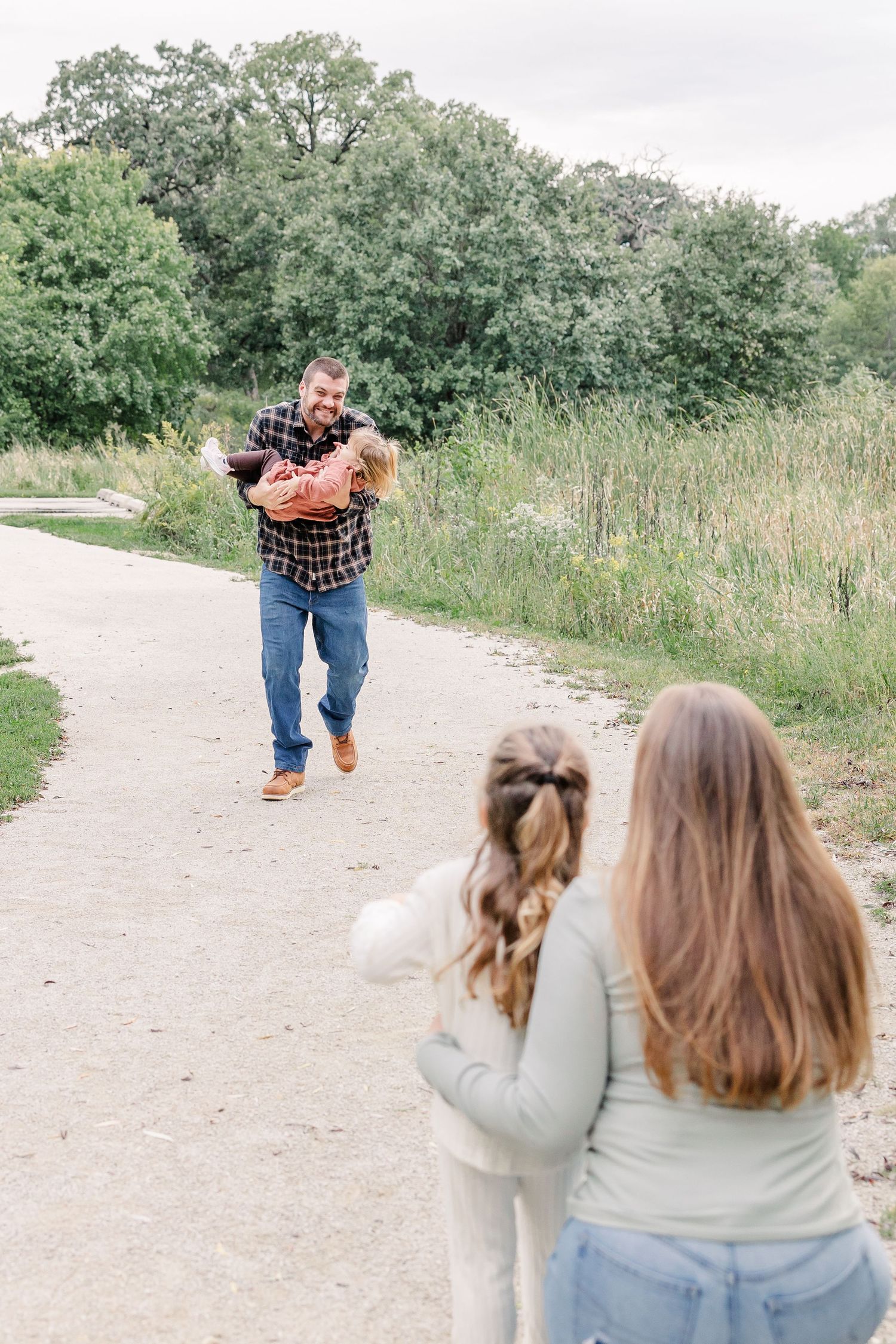 Smiling bearded dad playfully running toward mom and daughter on a winding nature path surrounded by lush green trees.