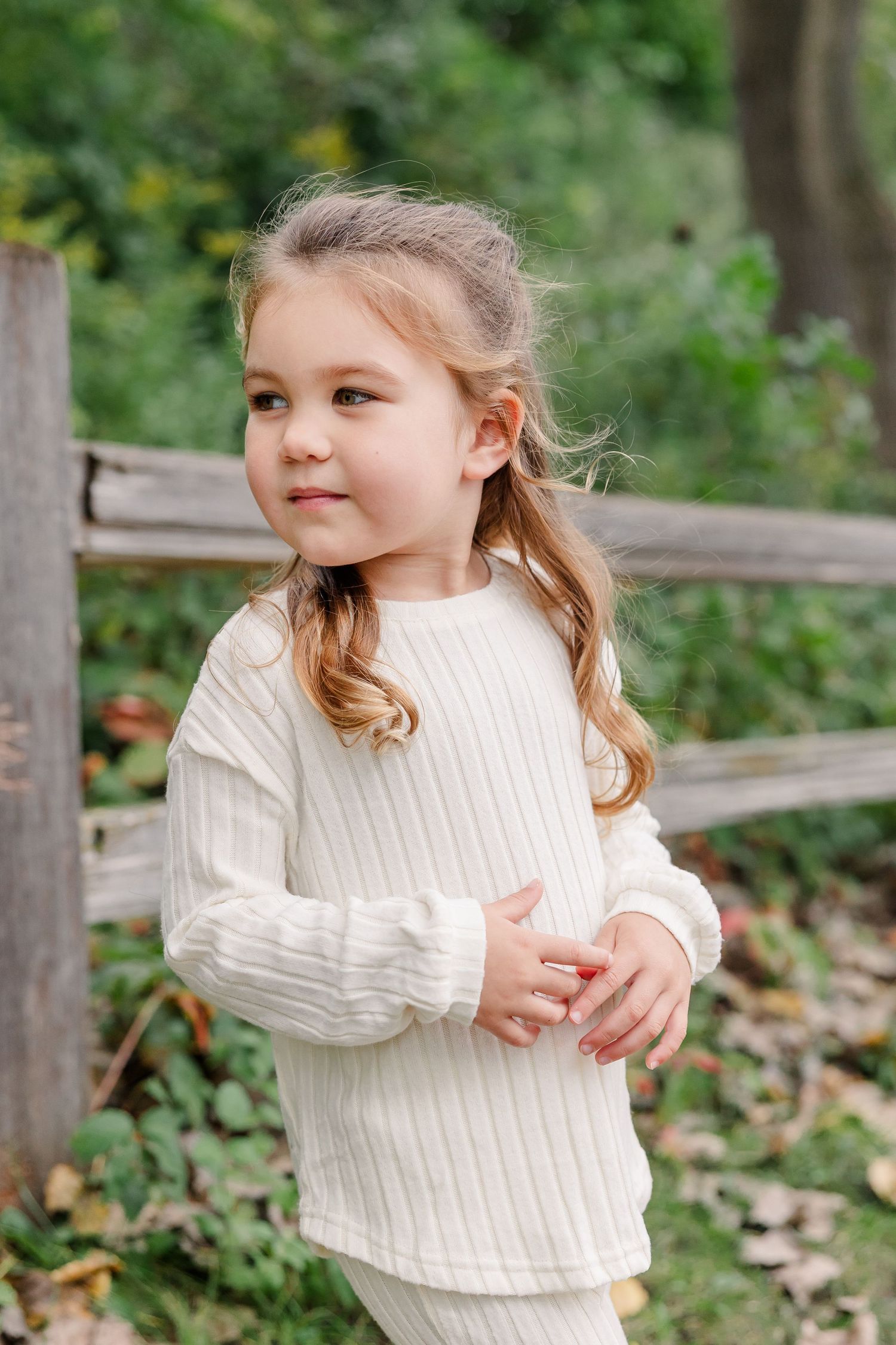 A young girl in a white ribbed sweater stands near a wooden fence in a lush green outdoor environment.