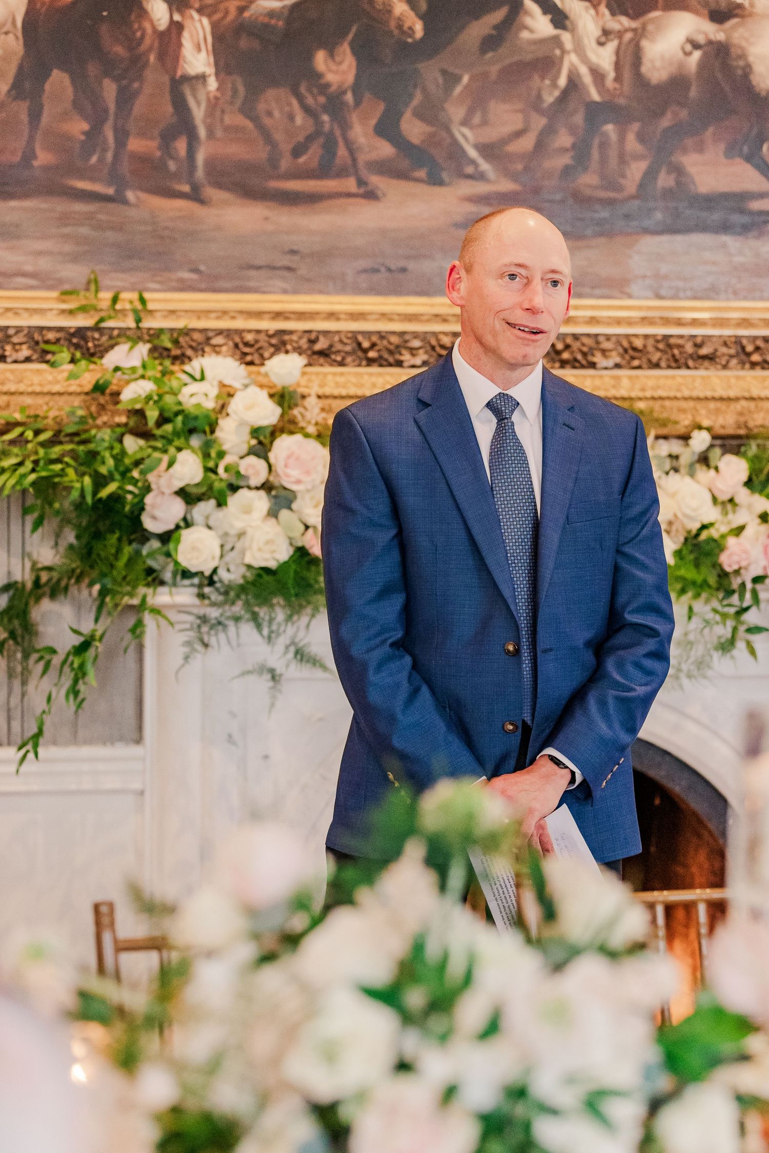 Guest speaker stands before equestrian painting and floral arrangements at formal wedding reception.