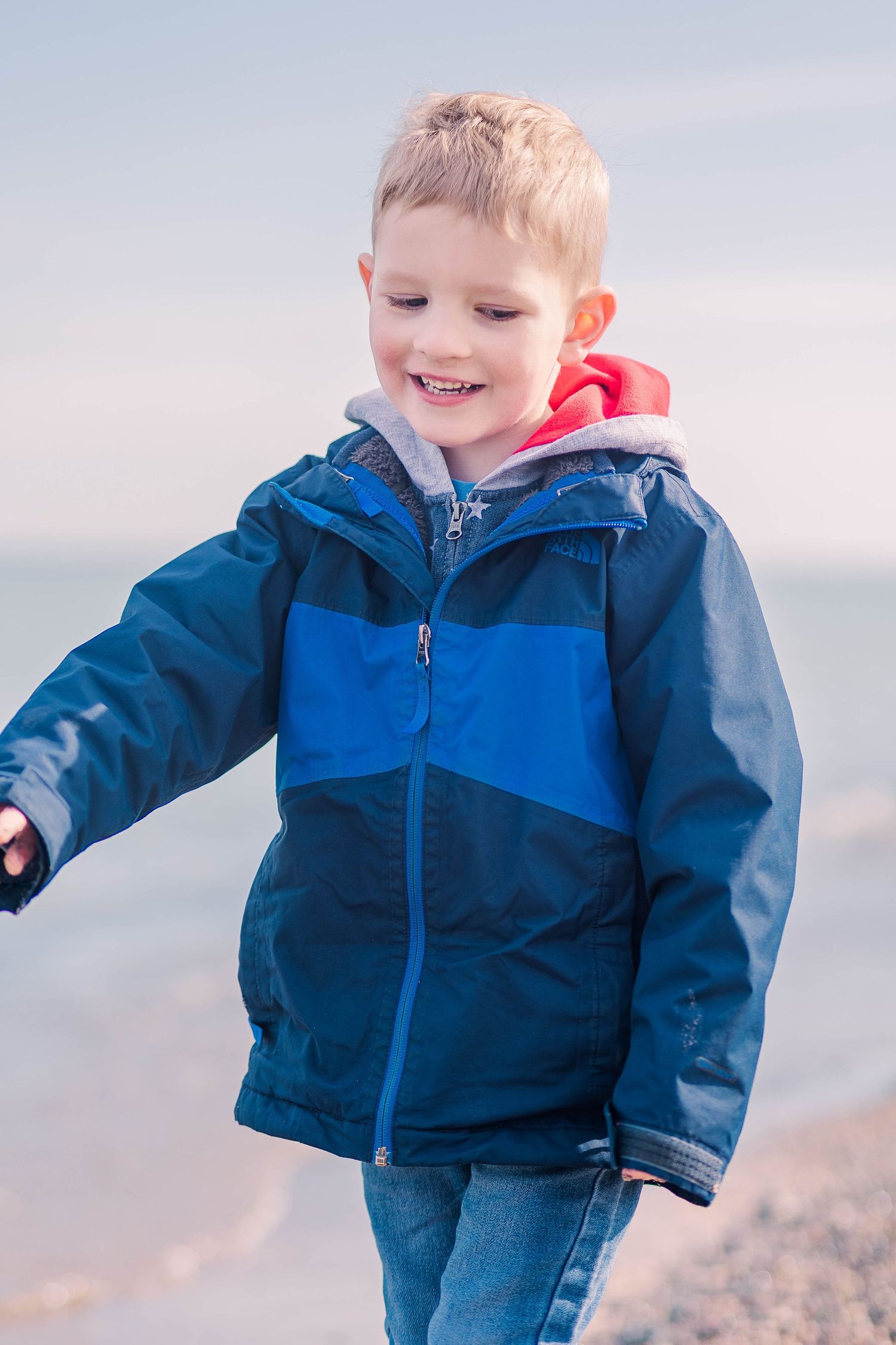 Child wearing navy and blue windbreaker smiles while exploring rocky shoreline.