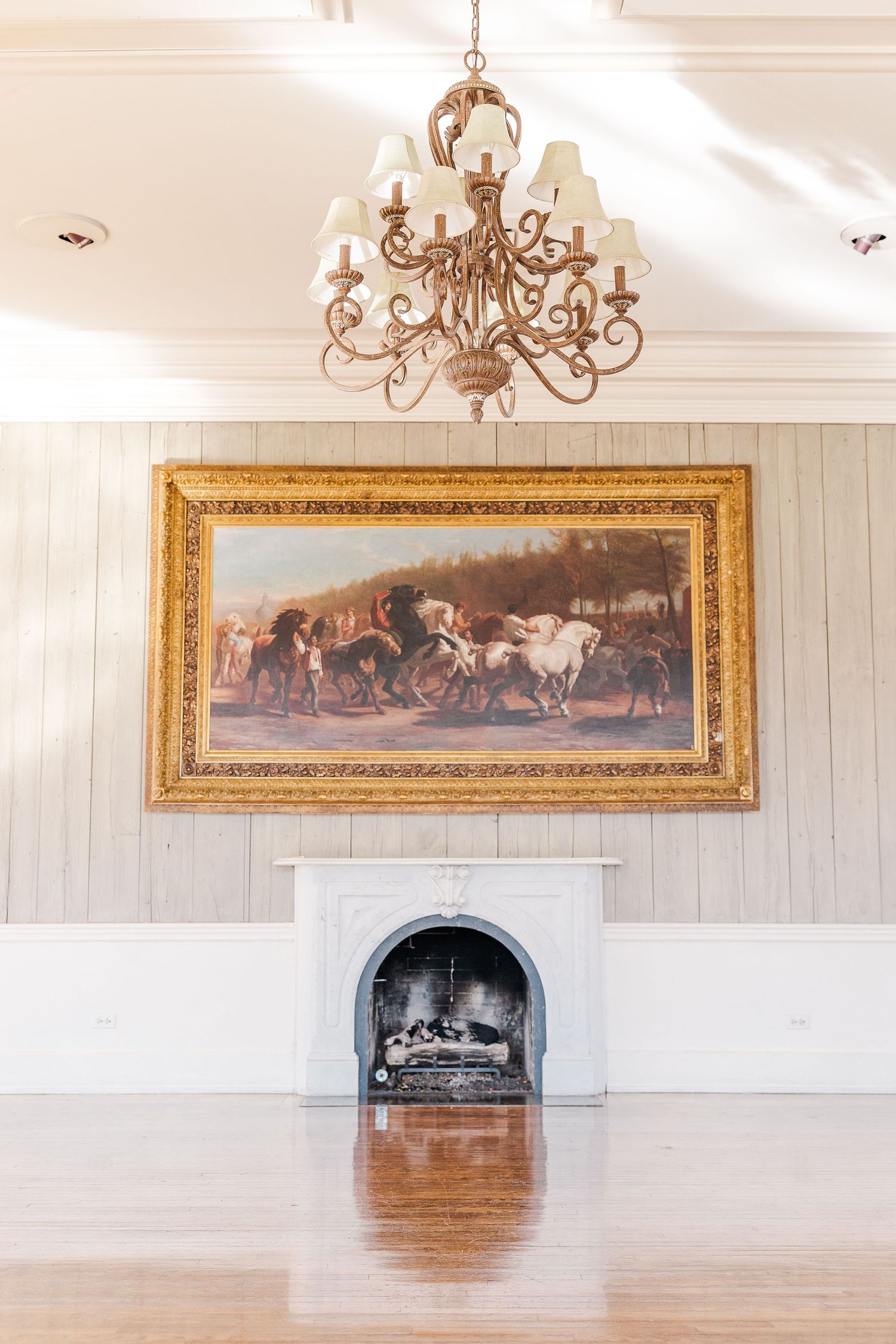 An elegant chandelier hangs above a framed horse painting and marble fireplace in a formal room.