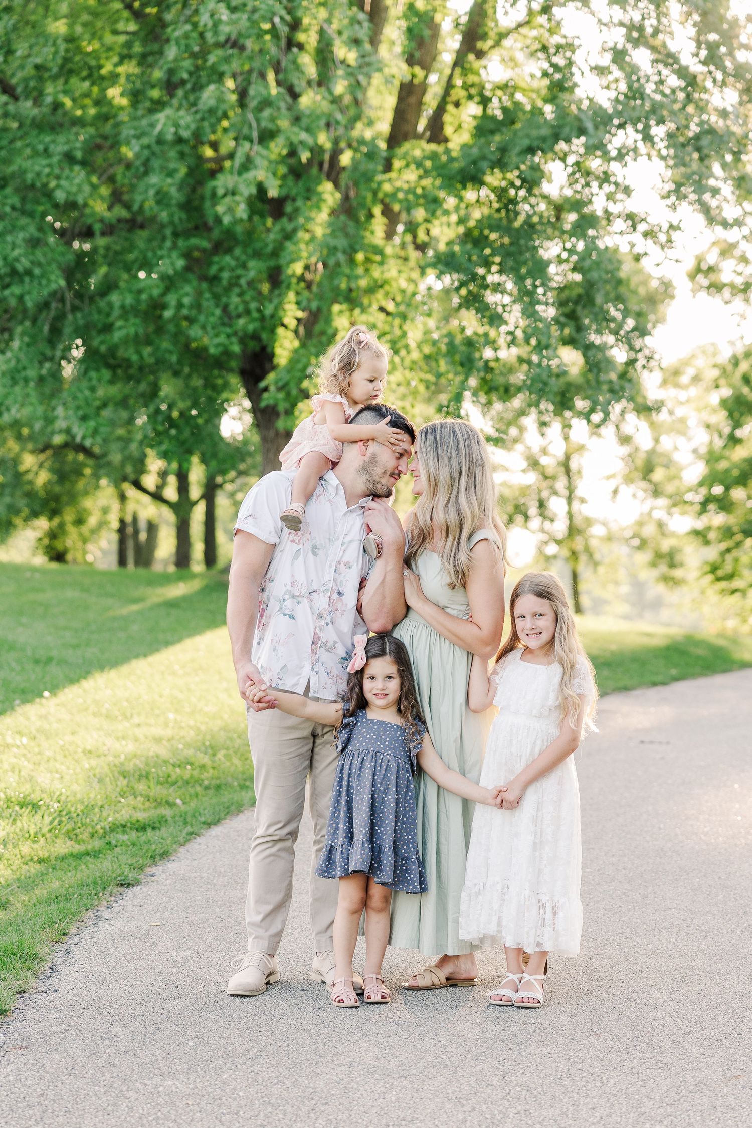 A family stands close together on a sunlit path with lush green trees in the background.