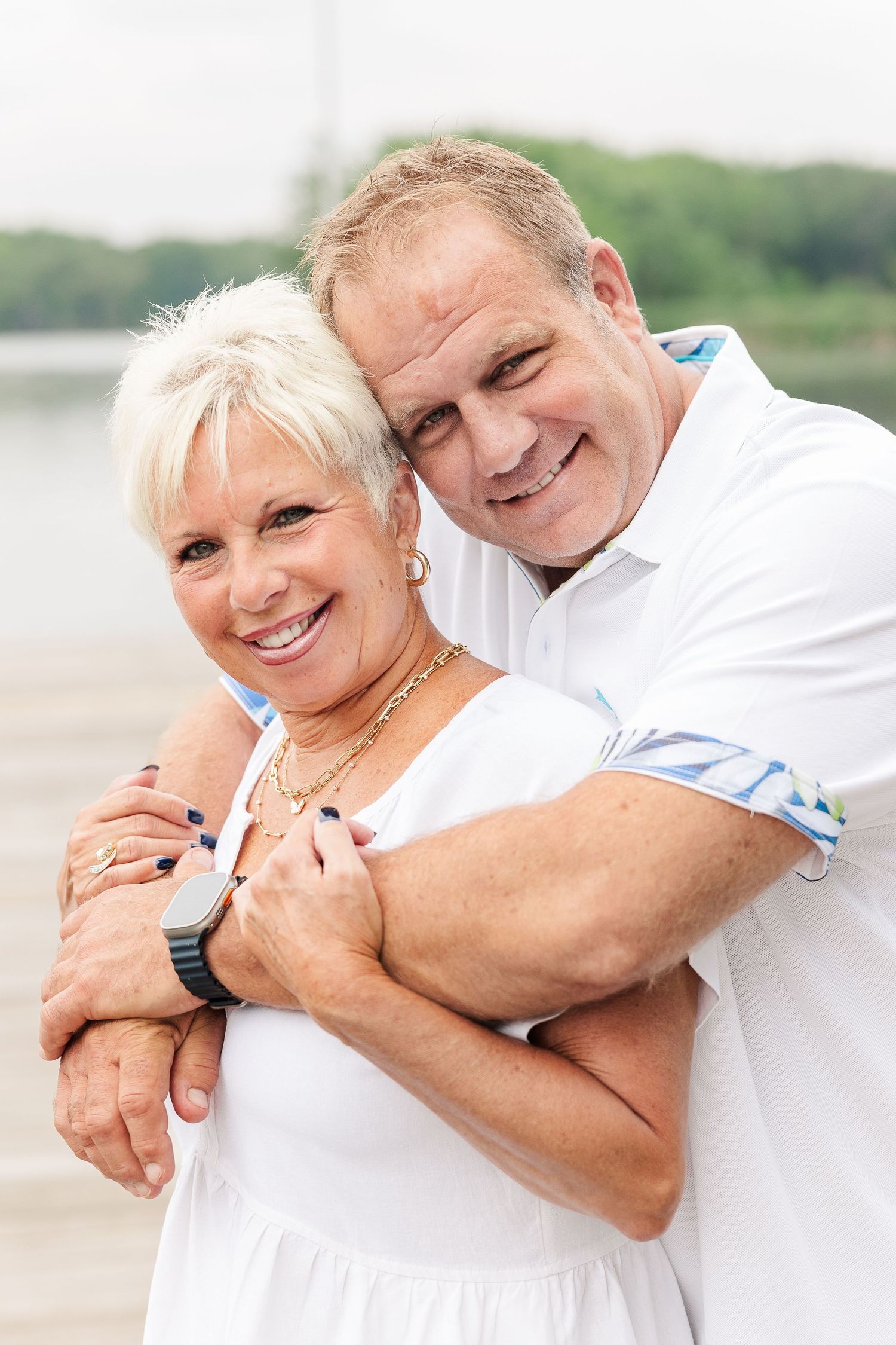 Couple in white outfits embrace while sharing joyful moment on dock by lake.